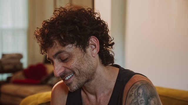 Man with curly hair smiling, sitting indoors with a blurred background.