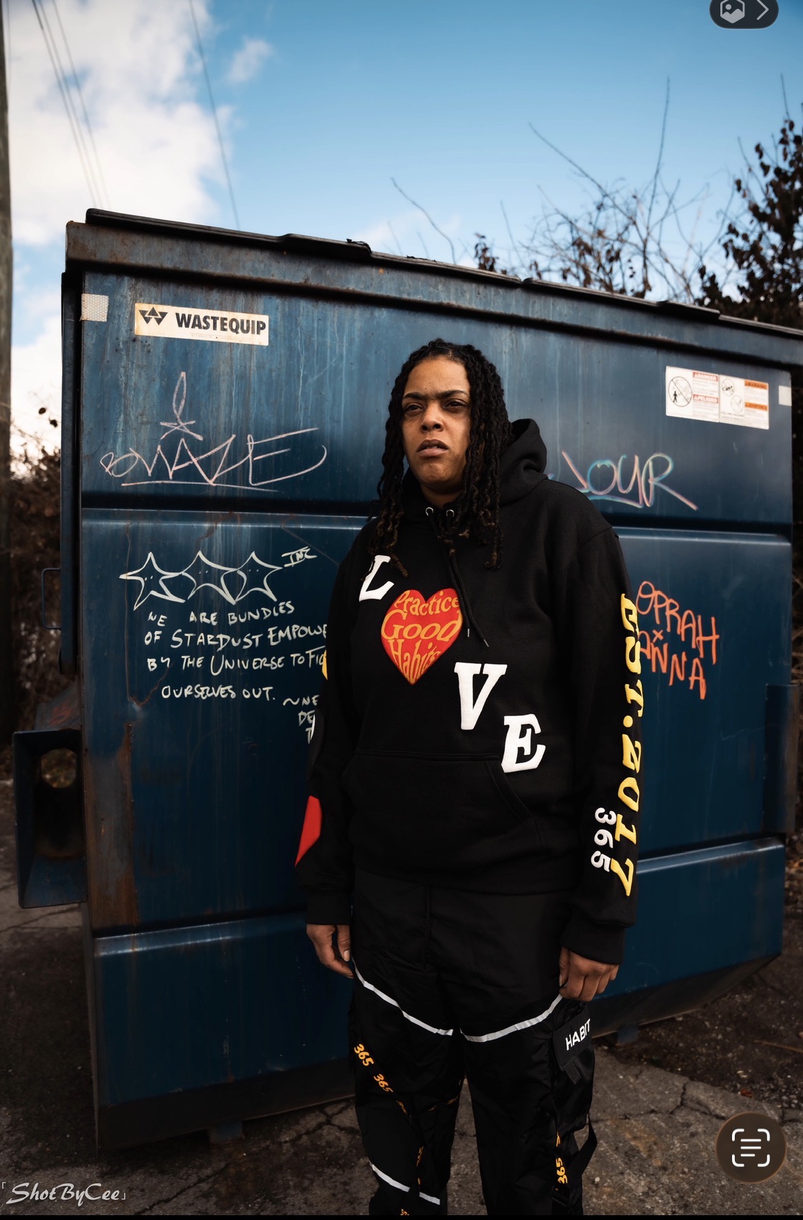 Person with long curly hair wearing a black hoodie with red and yellow text, standing in front of a blue dumpster.