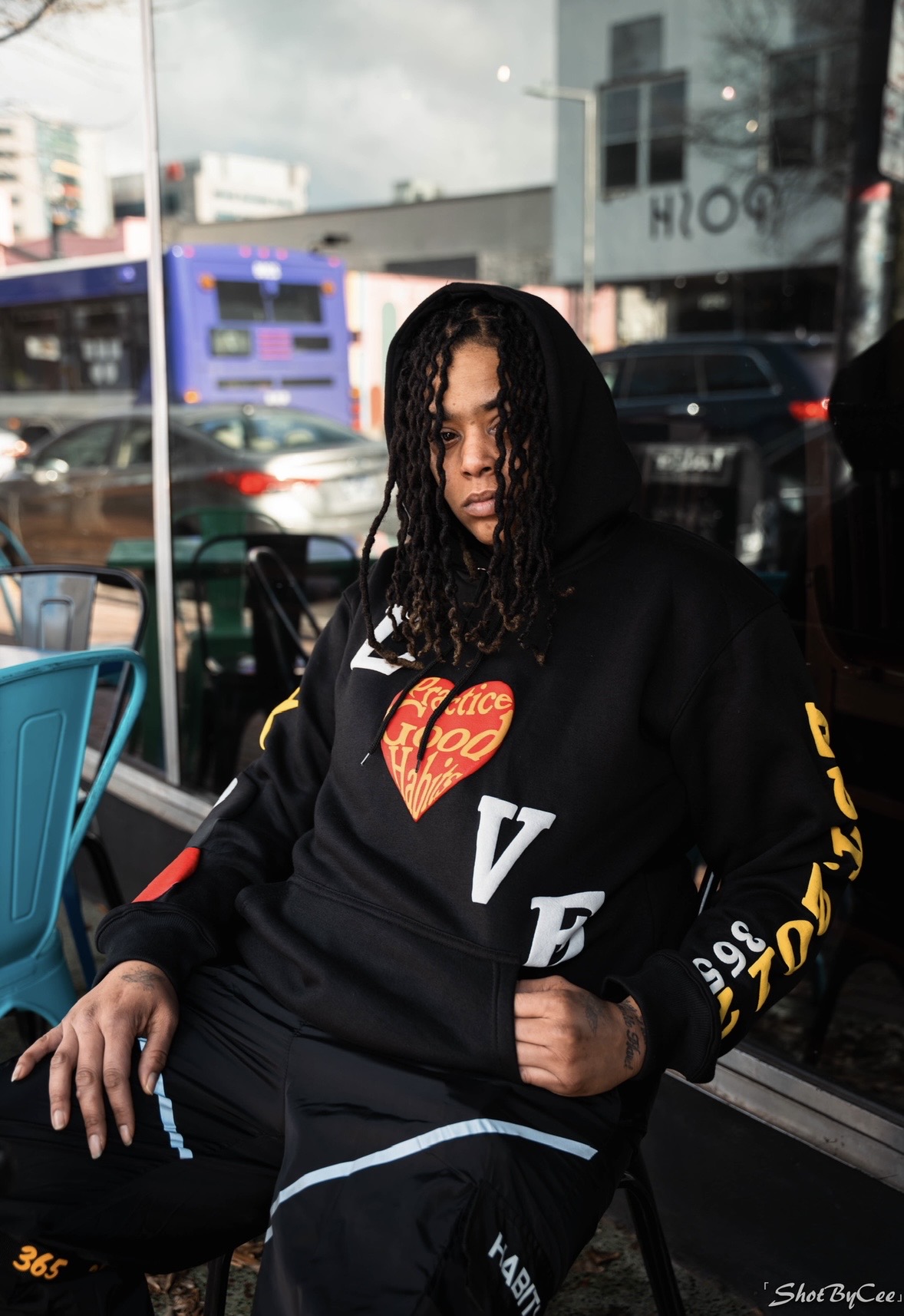 Young woman with curly hair wearing a black hoodie with colorful text, sitting outdoors near a glass window, with city street background.