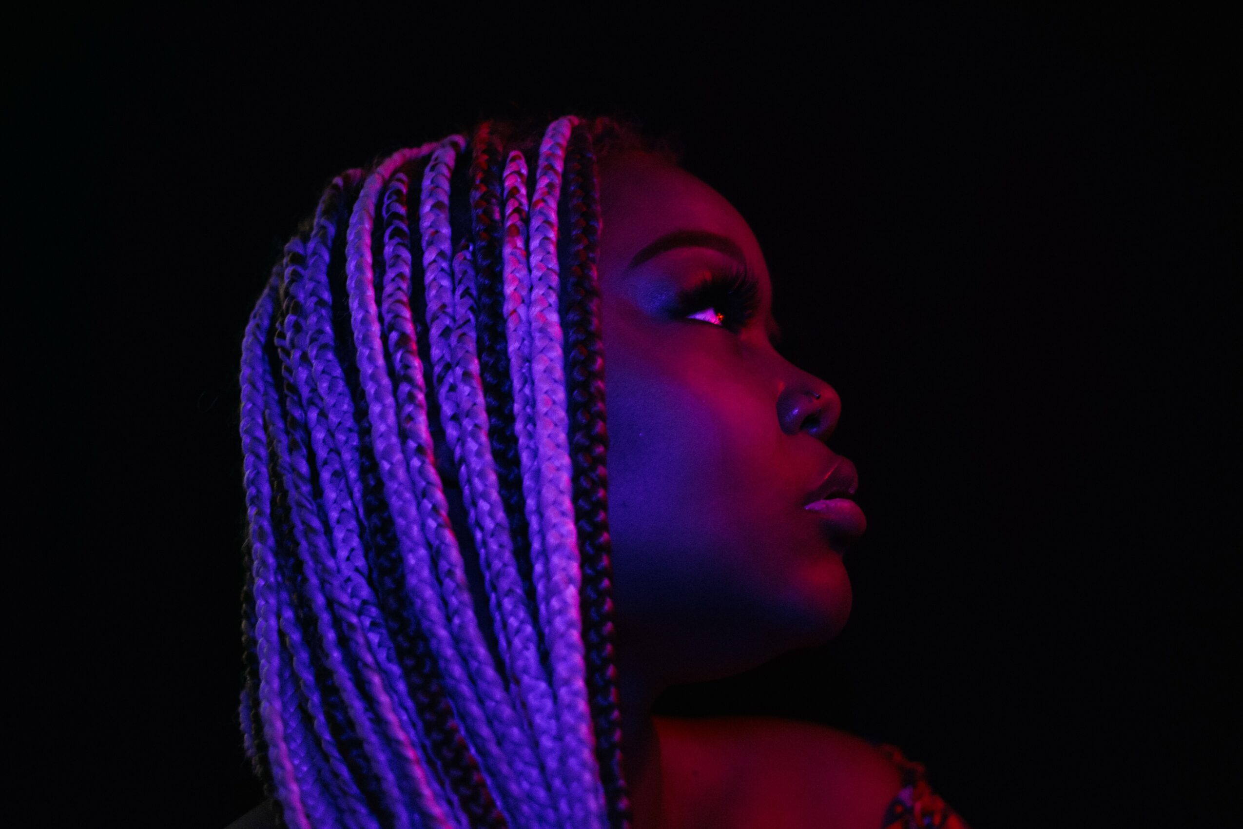Side profile of a woman with braided hair illuminated by purple and red lighting against a black background.