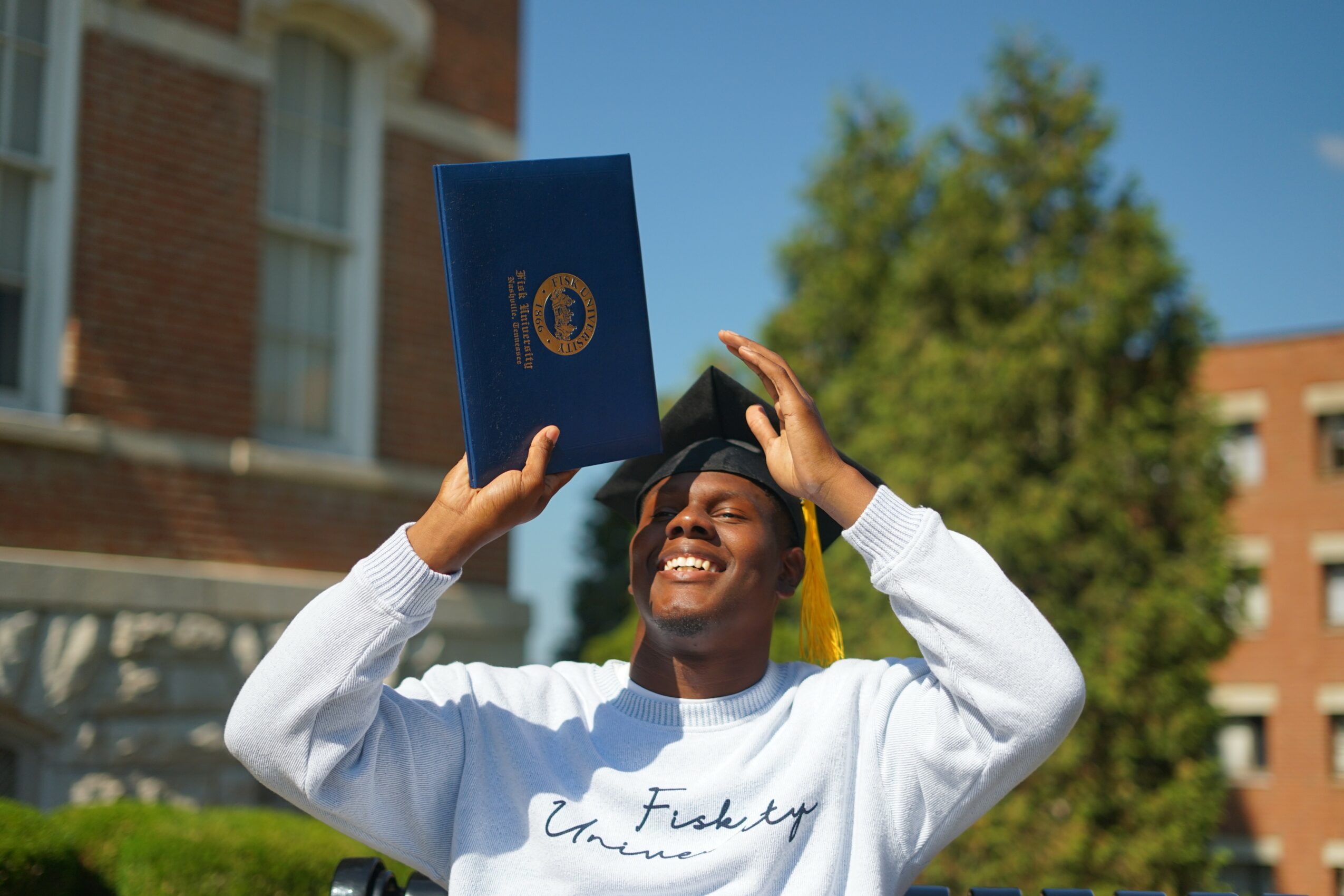 Graduate in cap and gown holding diploma outdoors, smiling, with trees and buildings in background.