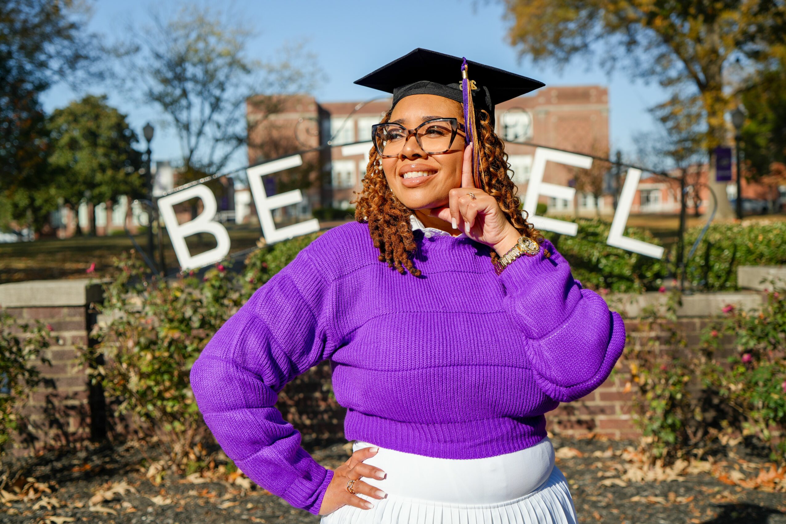 Woman in purple sweater wearing graduation cap, smiling outdoors with