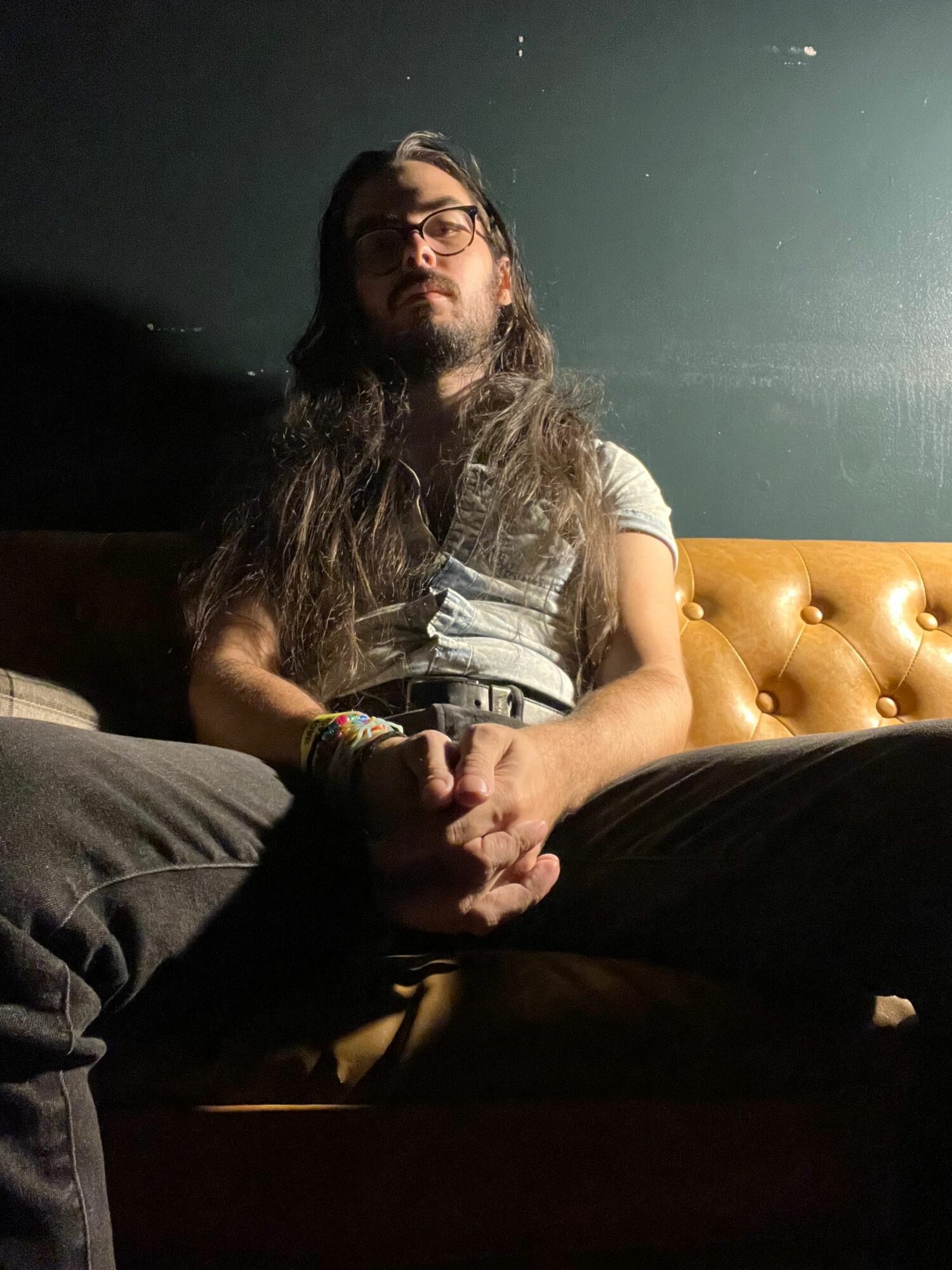 Person with long hair lying on a bed with a tufted headboard, holding hands with another person, in a dimly lit room.