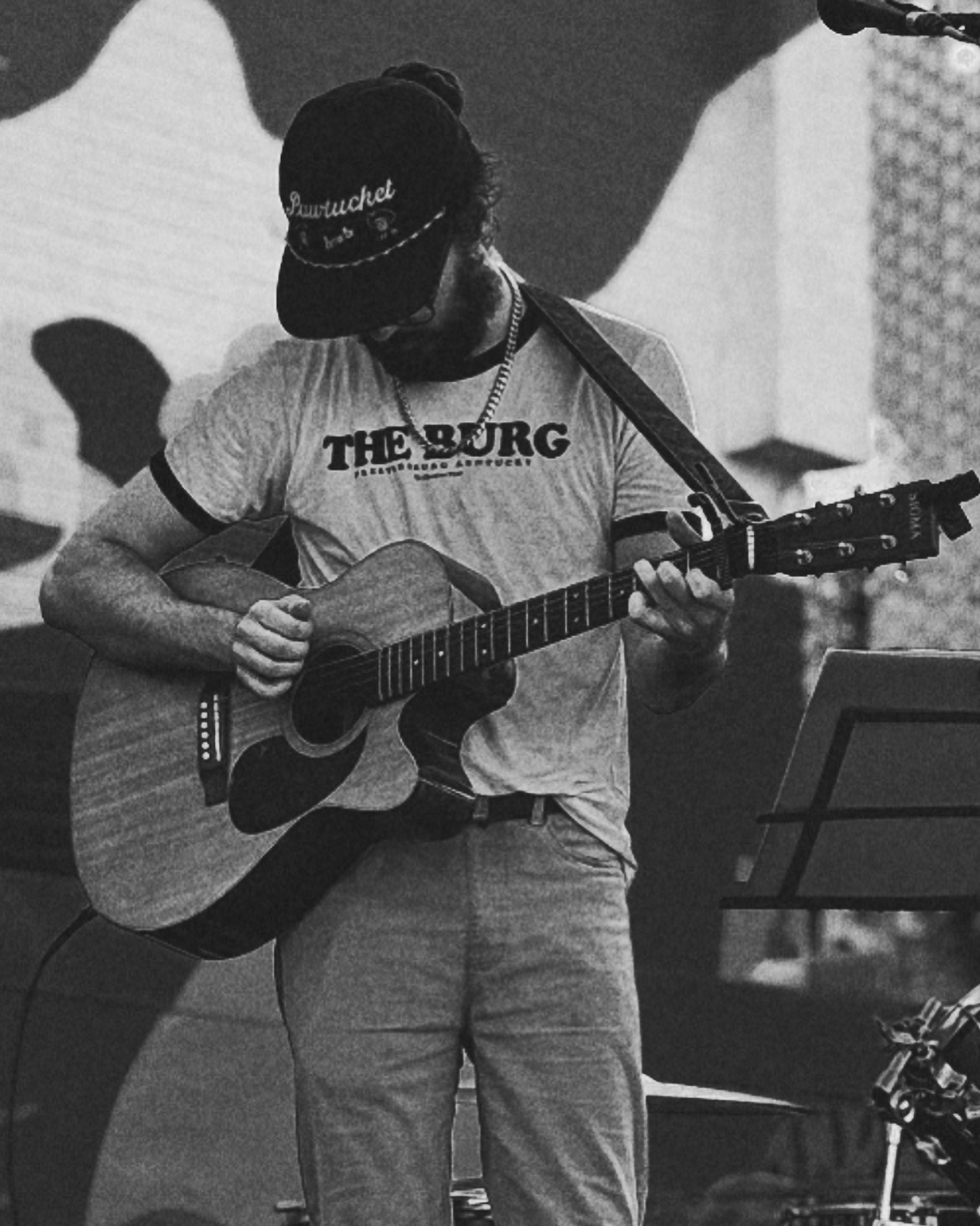 Person playing an acoustic guitar, wearing a cap and T-shirt, standing in front of a backdrop, with music stand nearby.
