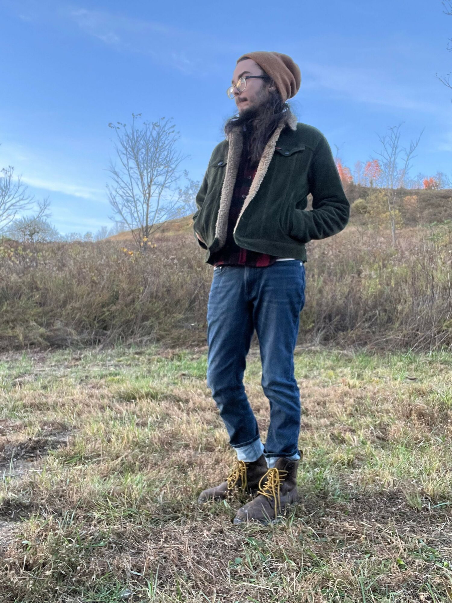 Person standing outdoors on grassy field with trees and cloudy sky in background, wearing a black jacket, jeans, and boots.