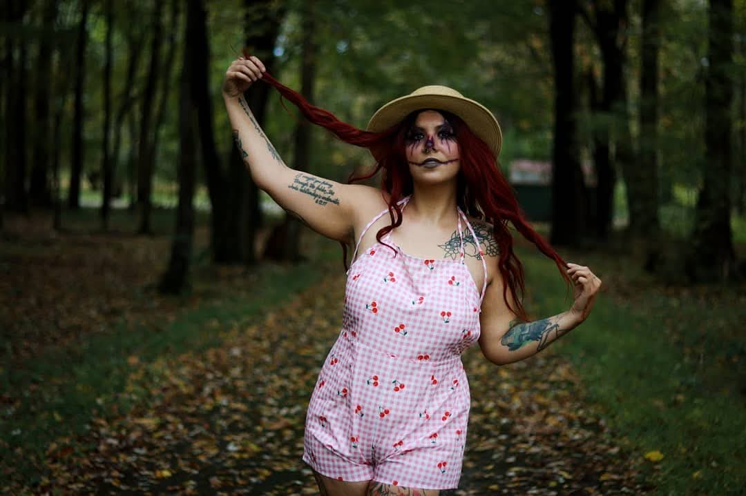 Woman with long red hair, wearing a hat and pink dress, standing in a forested park holding her hair.