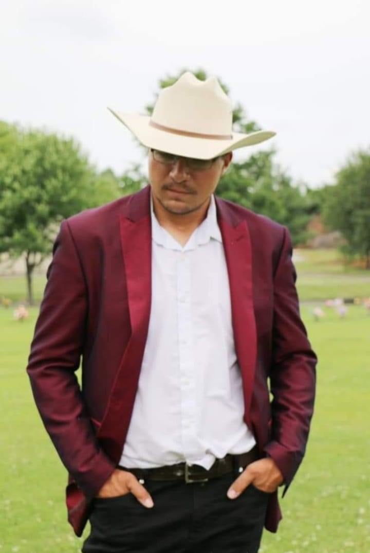 Man wearing a white hat, white shirt, and maroon blazer outdoors with trees and grass in background.