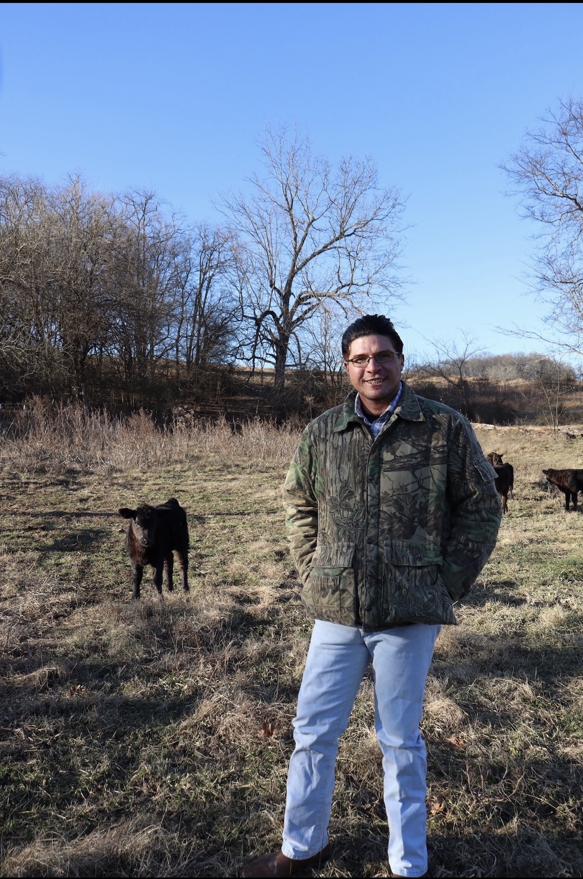 Man standing outdoors in a field with cows, trees, and a clear blue sky in the background.