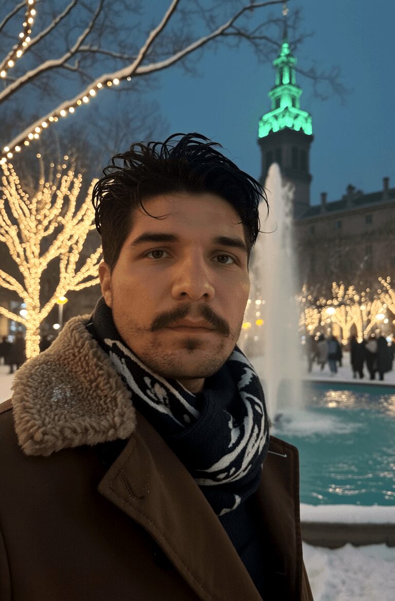 Young man with dark hair and mustache in winter coat and scarf, city fountain, illuminated trees, and a tower in background.