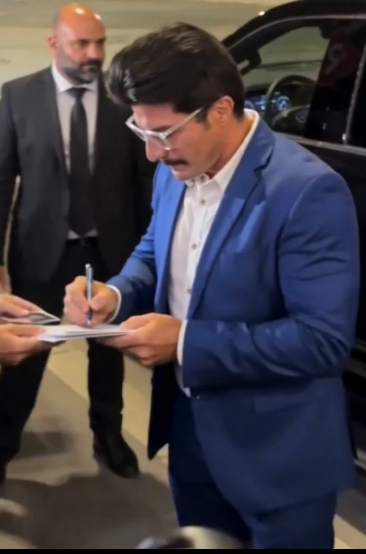 Man in blue suit signing a document, with another man in a suit in the background, in an indoor setting.