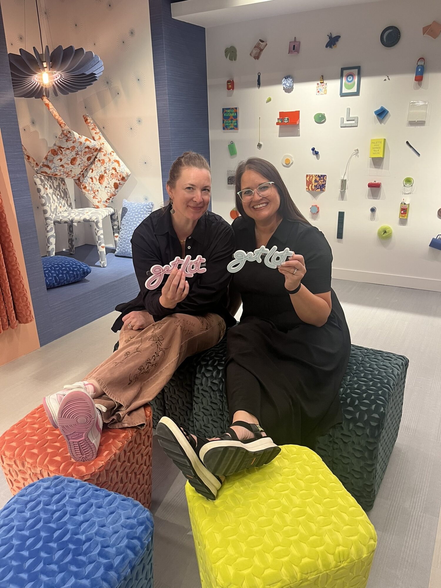 Two women sitting on a textured bench, smiling and holding signs, in a colorful, decorated indoor space.
