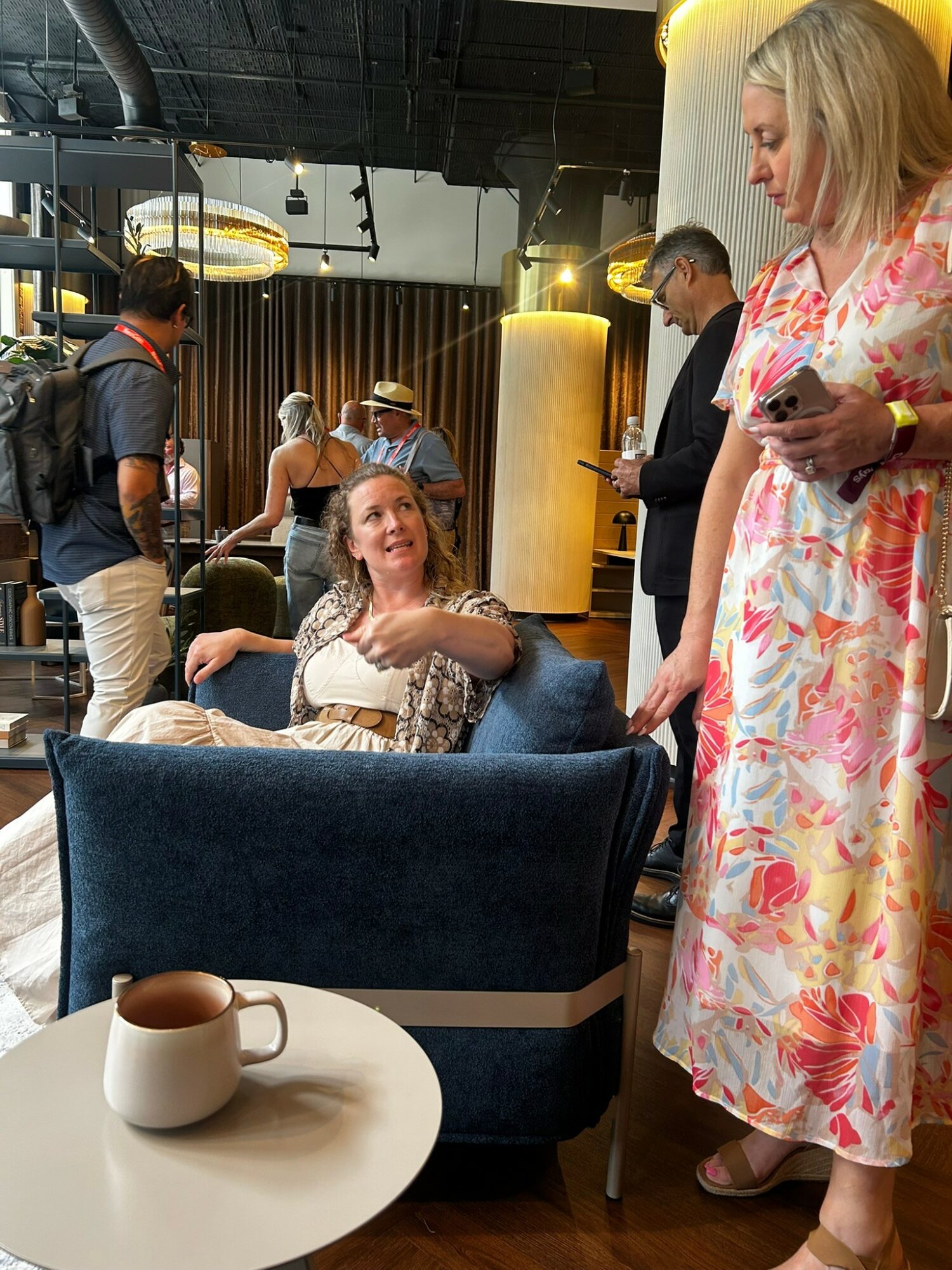 Woman sitting on a blue armchair, smiling and talking to standing woman holding a phone, in a busy indoor setting.