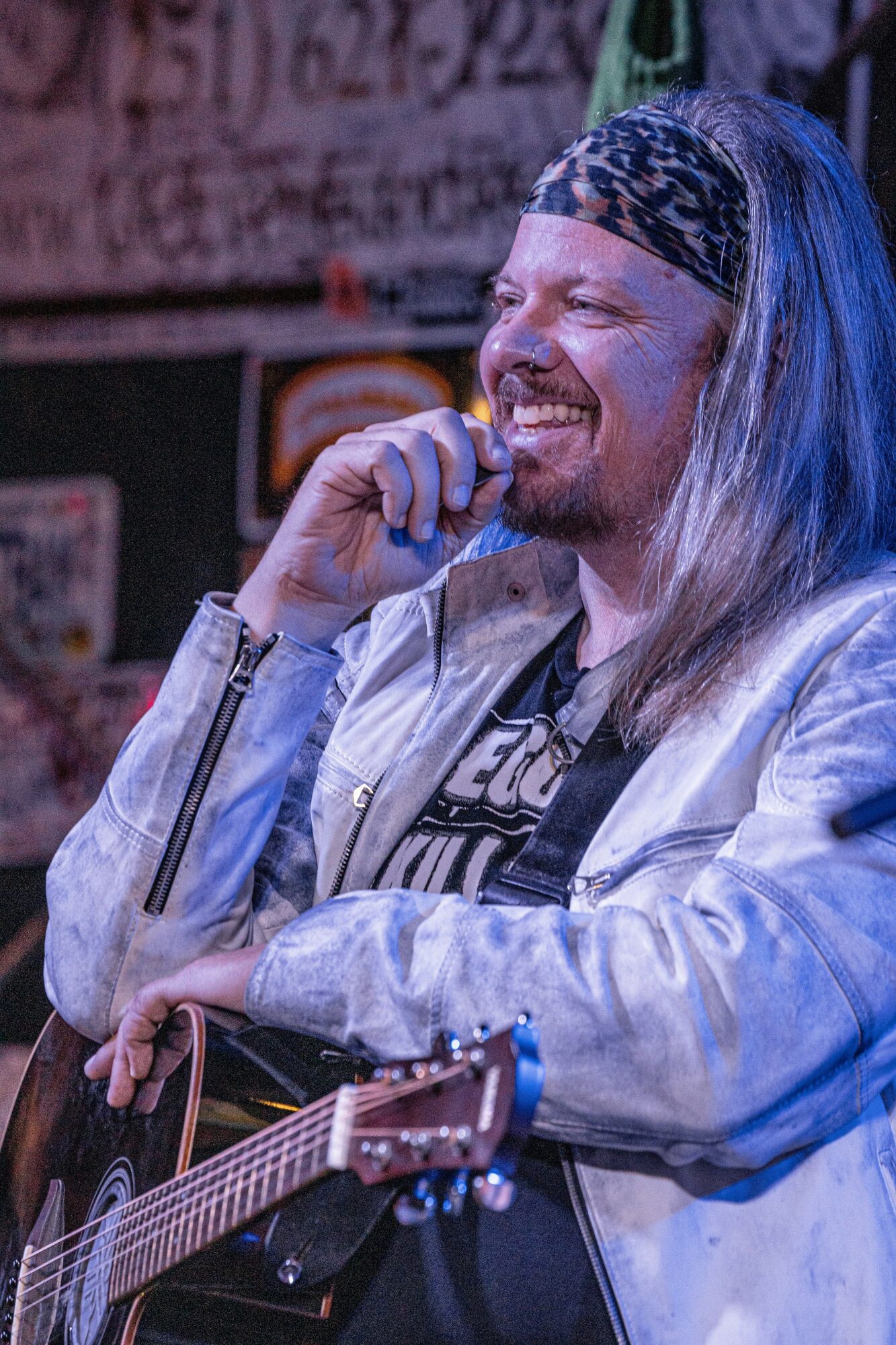Person with long hair, headband, smiling, holding a guitar, in a lively setting.