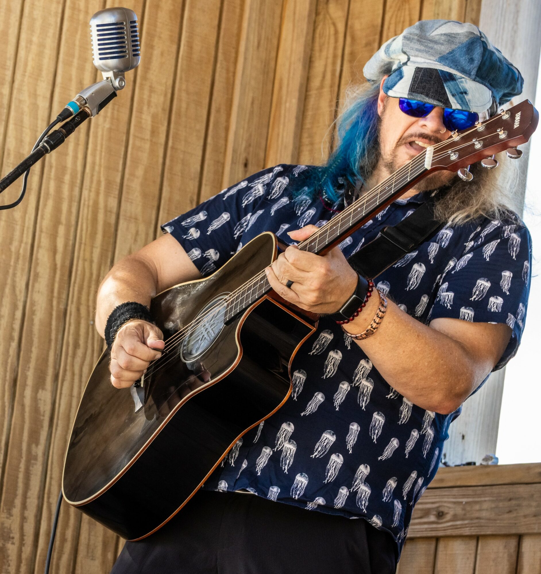 Person with blue hair, sunglasses, and a hat playing an acoustic guitar in front of a wooden wall.