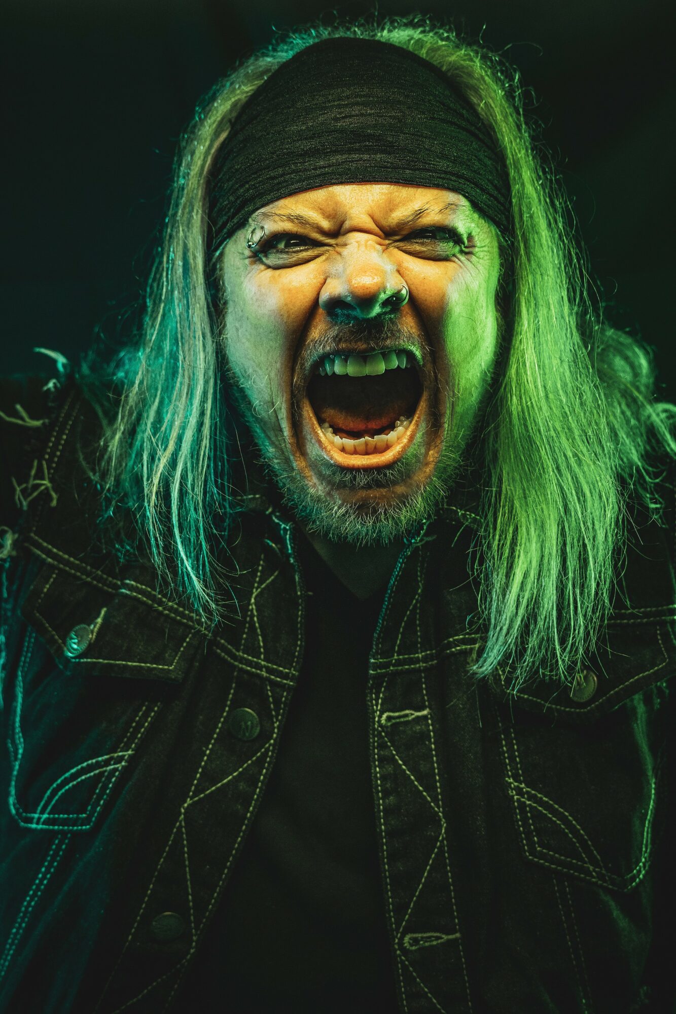 Person with long hair, headband, shouting or screaming, with intense facial expression, in dark lighting with green highlights.