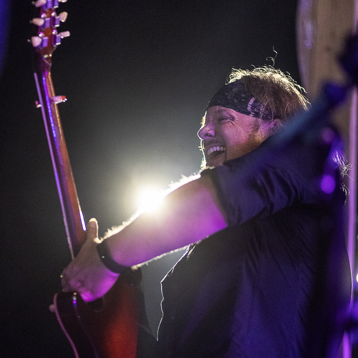 Person smiling and playing guitar on stage with bright backlight, wearing a headband and dark clothing.