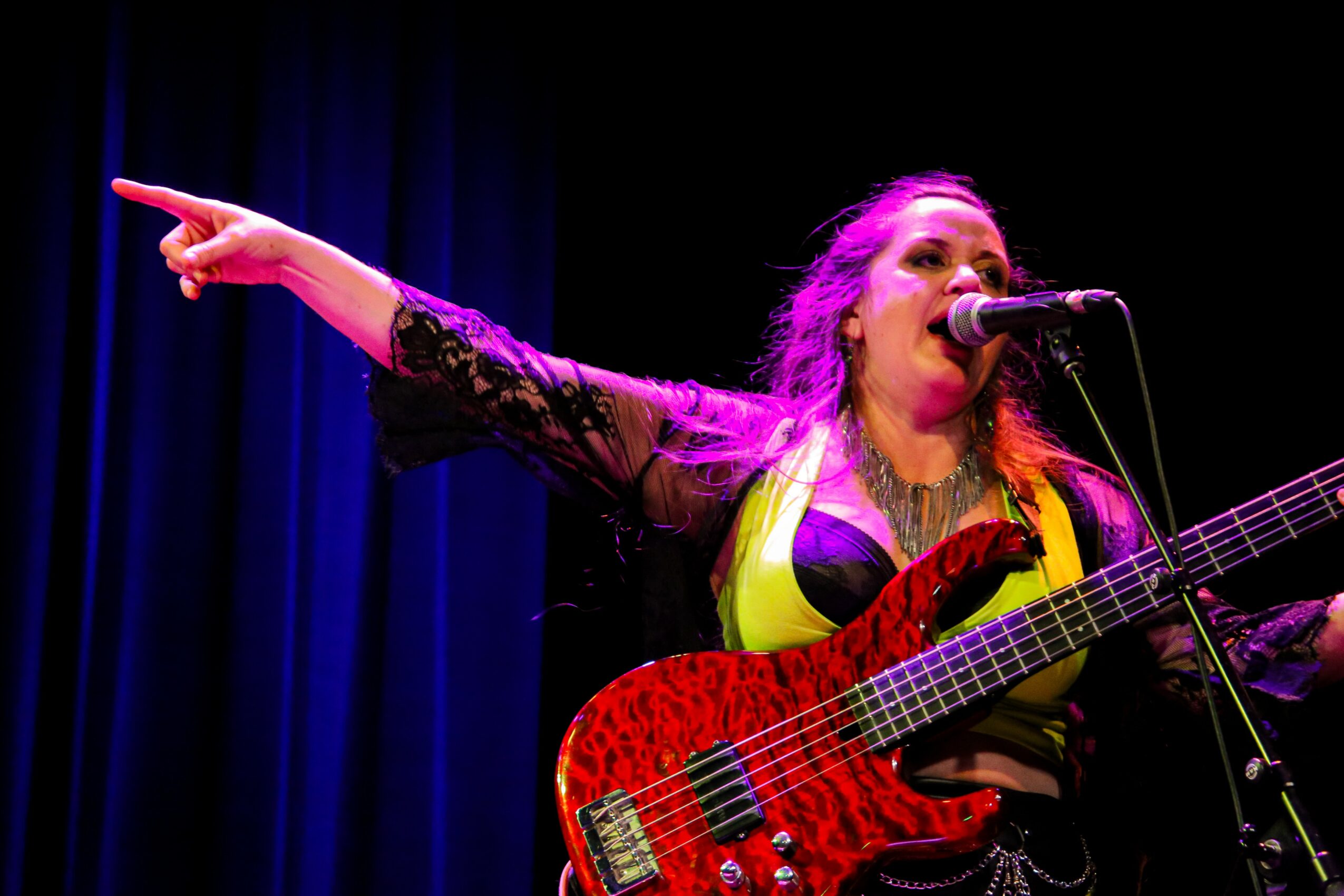 Female musician singing into a microphone and playing a red electric guitar on stage with a dark background.