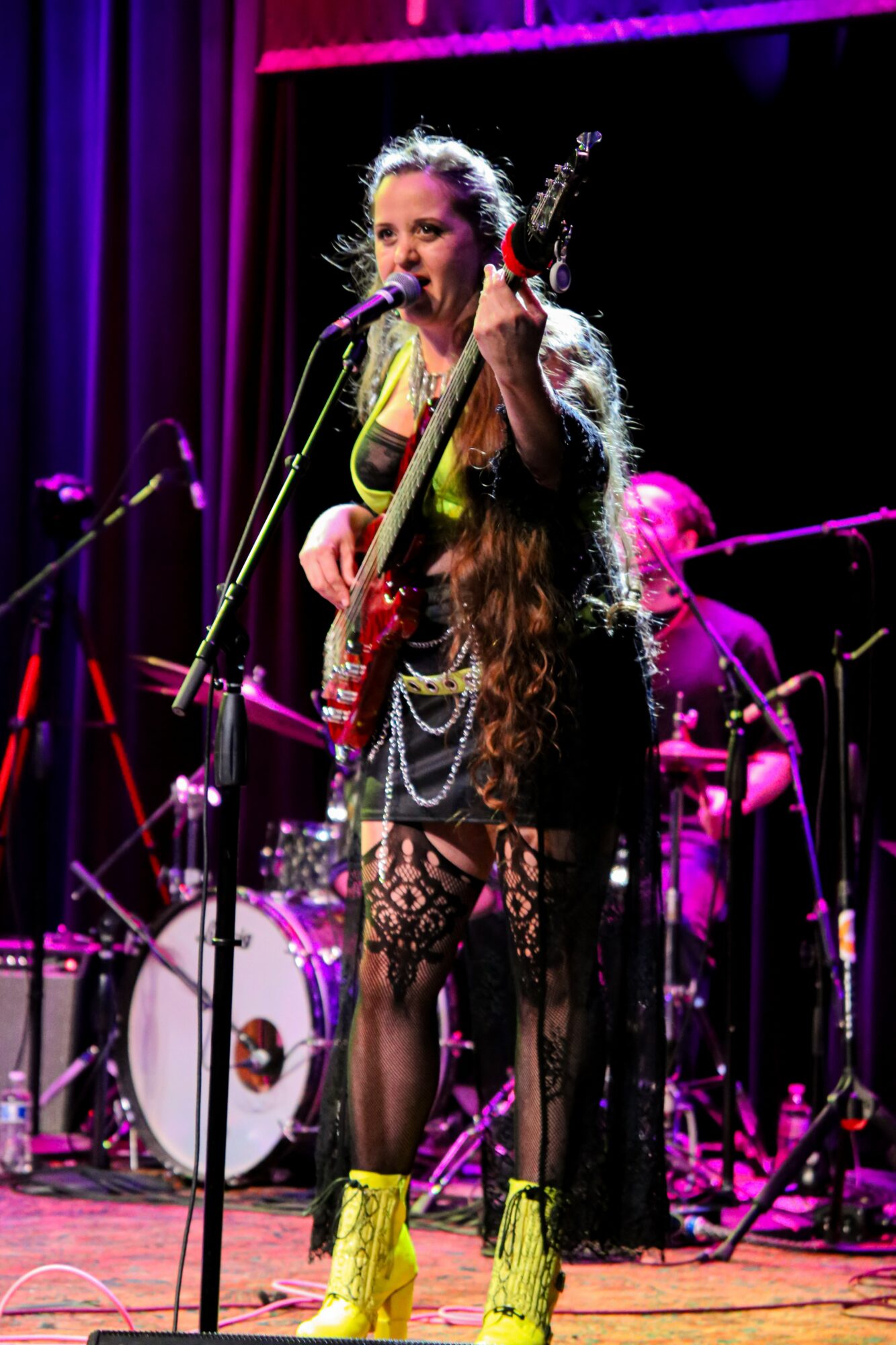 Female musician singing into microphone, playing guitar, on stage with drums in background, purple lighting, wearing yellow shoes and lace outfit.