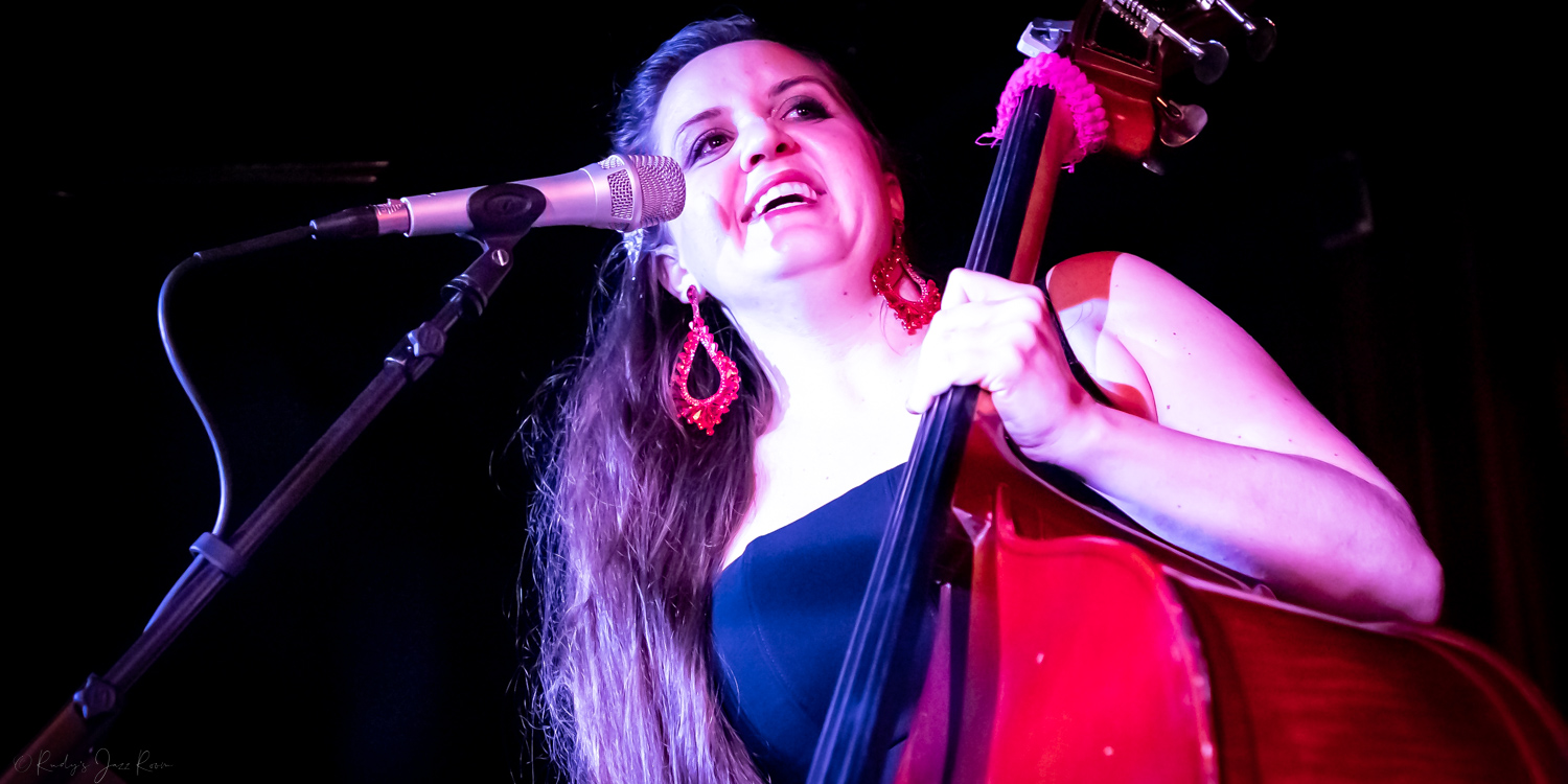 Woman singing into microphone while holding a red guitar, smiling, with dark background.