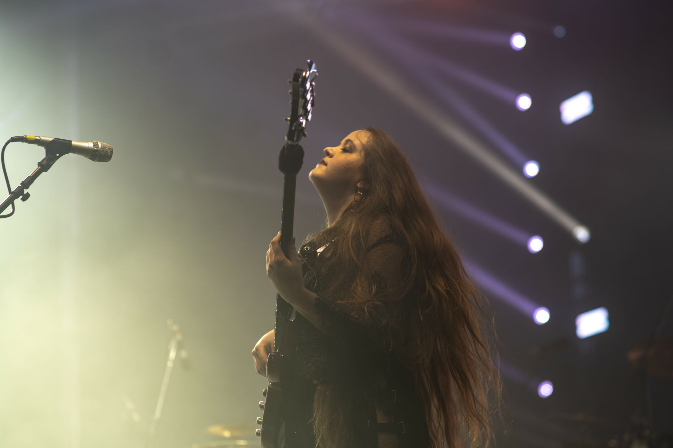 Woman playing guitar on stage with bright lights and fog, long hair flowing, looking upward.