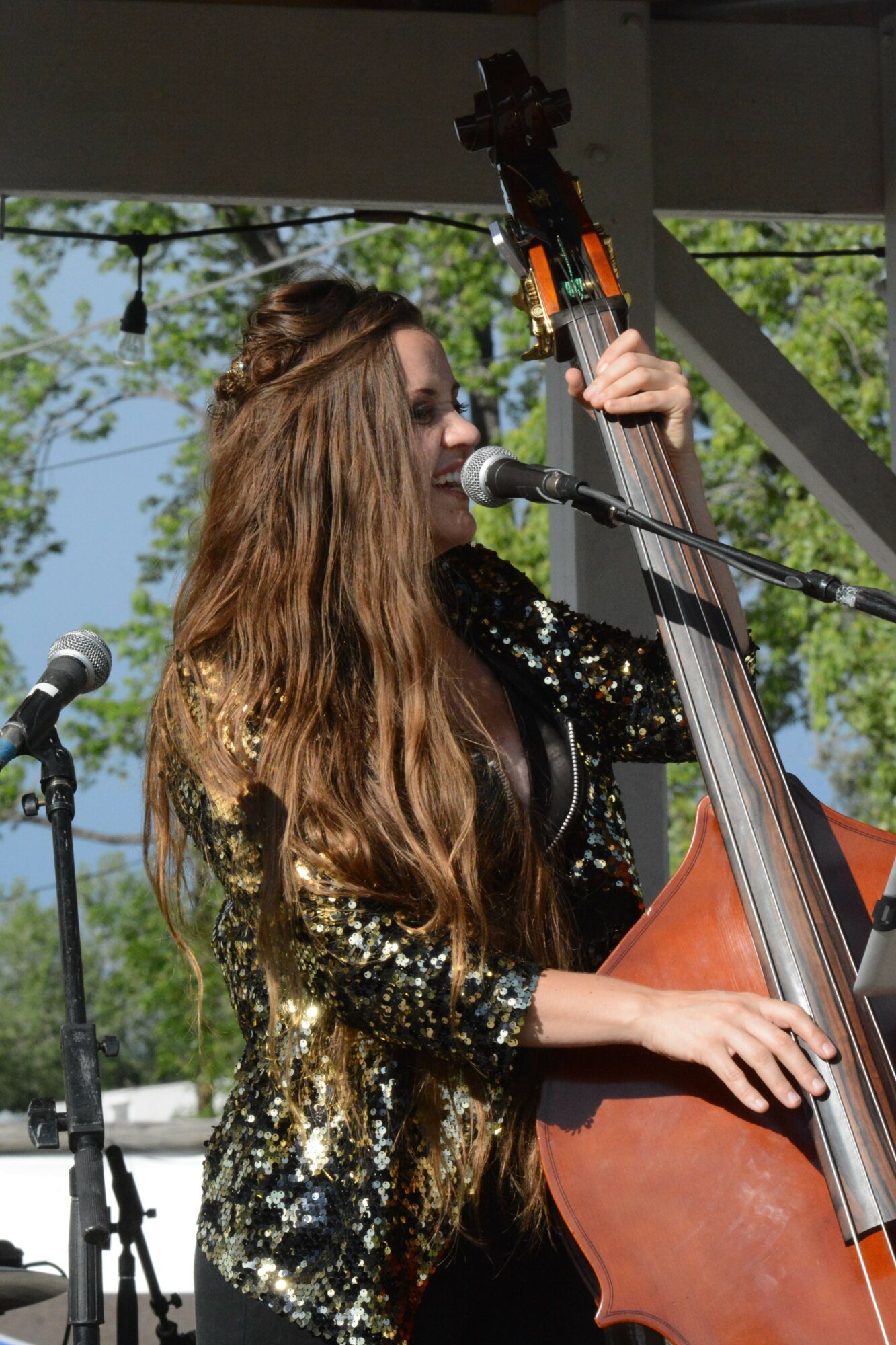 Person with long hair playing a string instrument outdoors, with trees and sky in background.