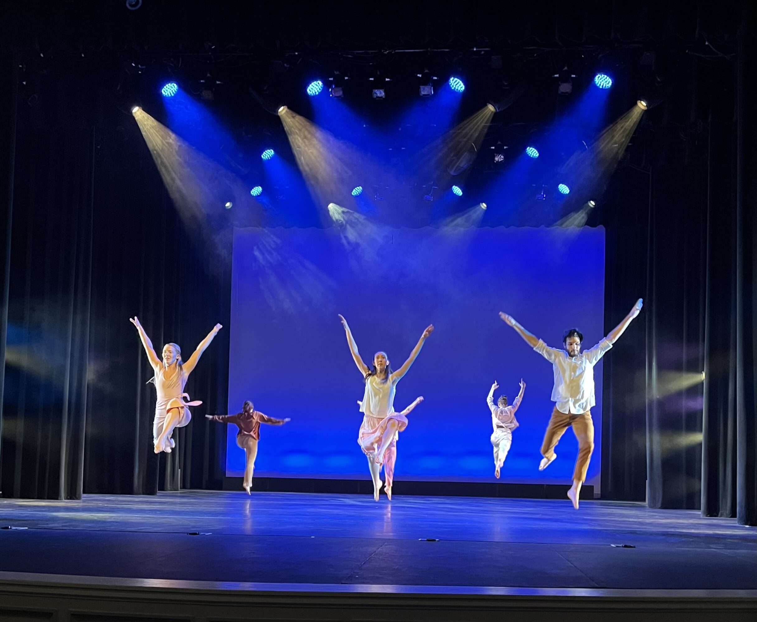 Six dancers perform on stage with colorful lighting and a blue backdrop, mid-air with arms raised, in a dance routine.