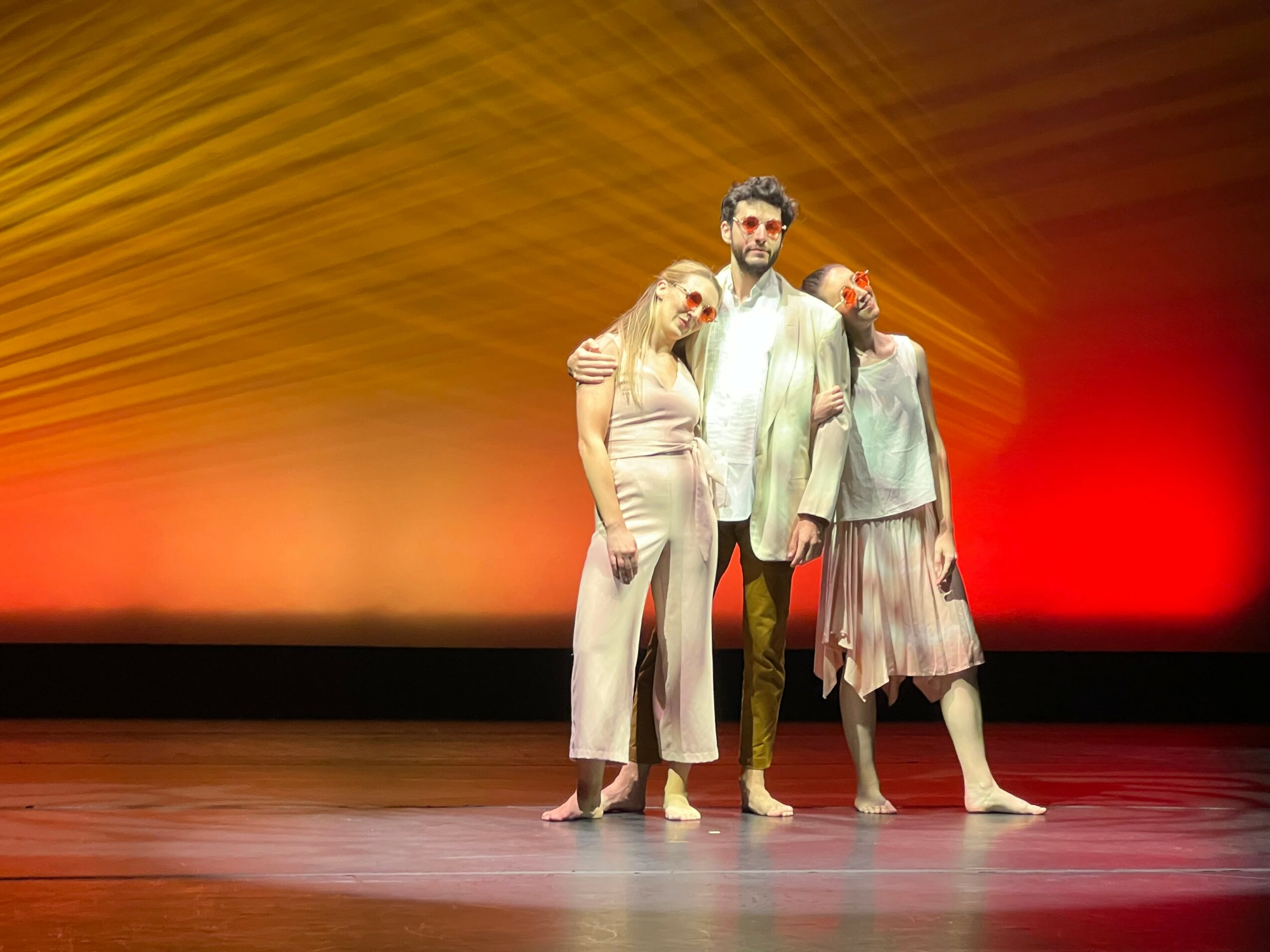 Three performers stand on stage with colorful background, two women leaning on a man in the center, all barefoot.