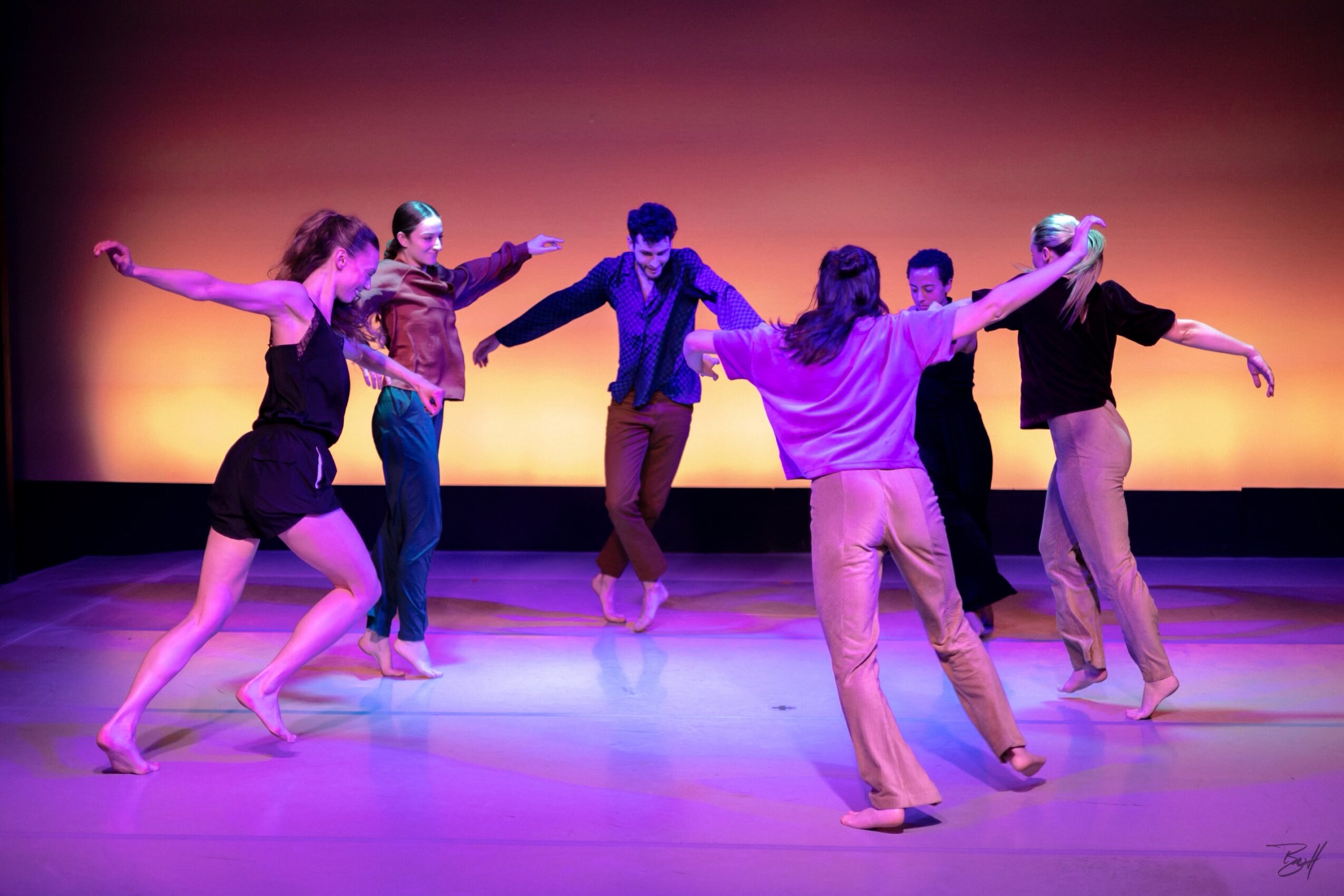 Group of seven dancers holding hands in a circle on stage with colorful lighting background.