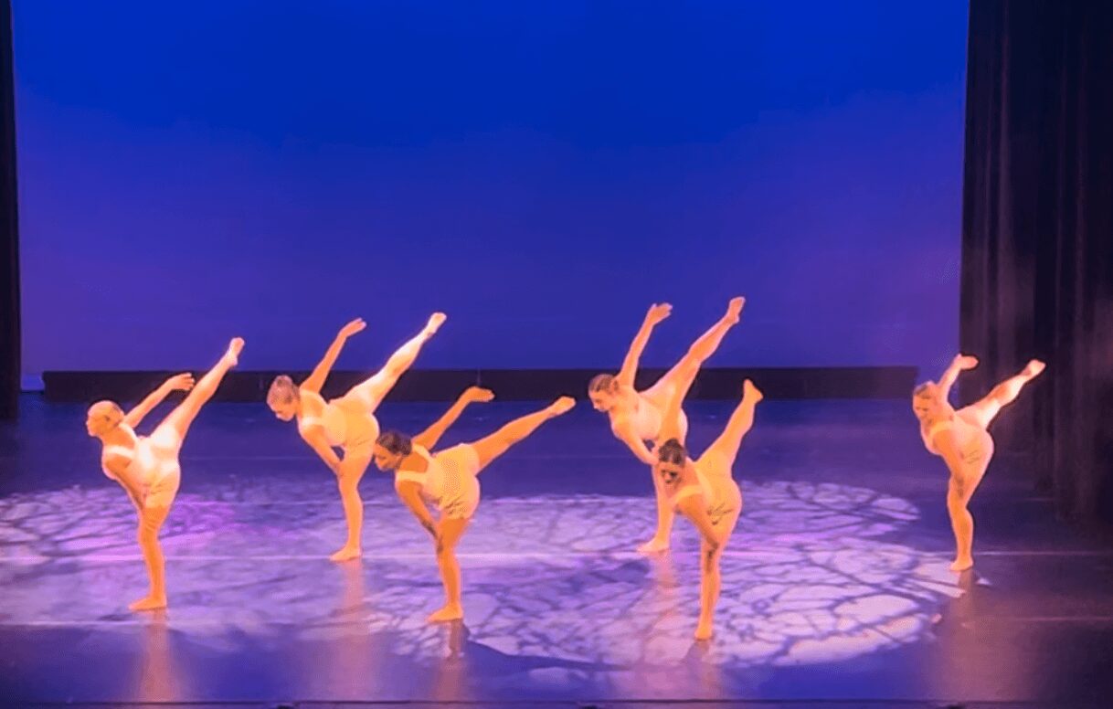 Five dancers perform a synchronized move on stage, each balancing on one leg with the other leg extended upward, arms reaching out.