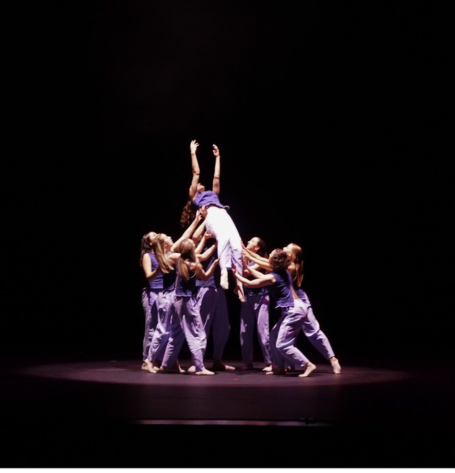 Group of children in purple costumes performing a dance move on stage, lifting a girl upside down.