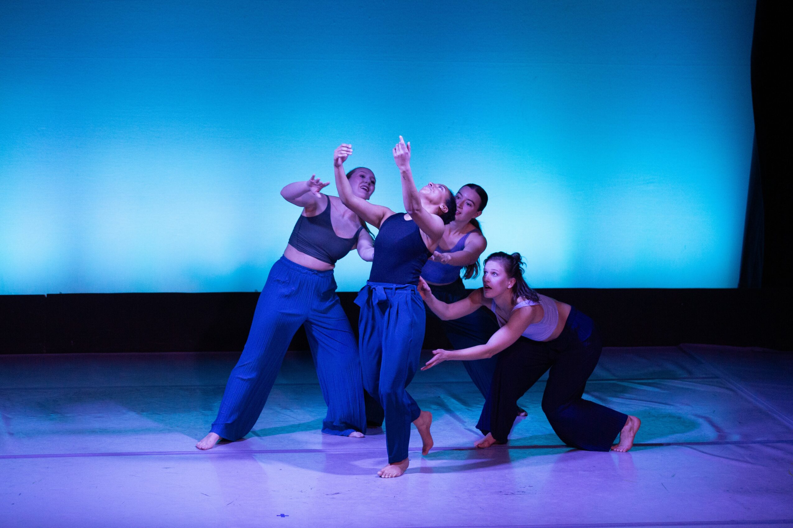 Four dancers perform on stage with a blue background, one crouching, others standing and gesturing, in a dance routine.