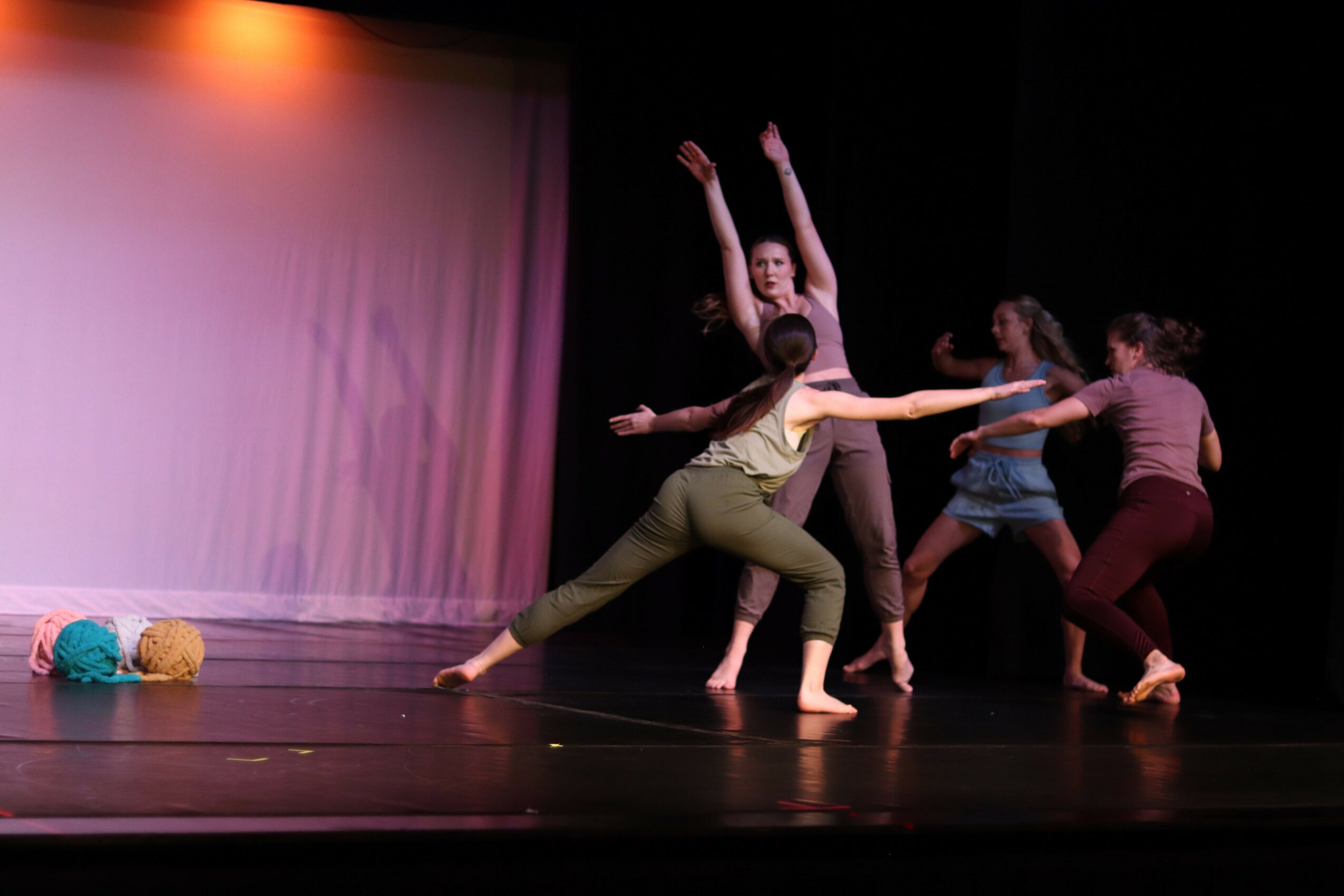 Group of five dancers performing on stage with colorful props, some dancers reaching upward, others in dynamic poses.