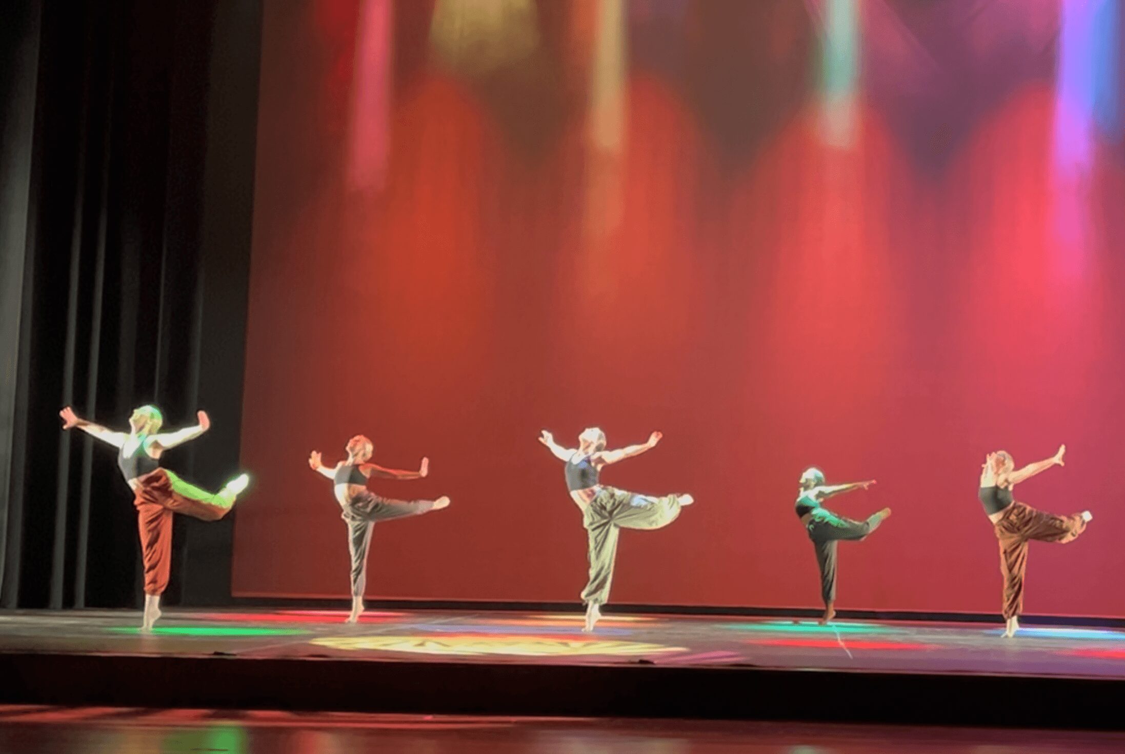 Six dancers perform on stage with colorful lighting and a plain backdrop, each balancing on one leg with arms extended.