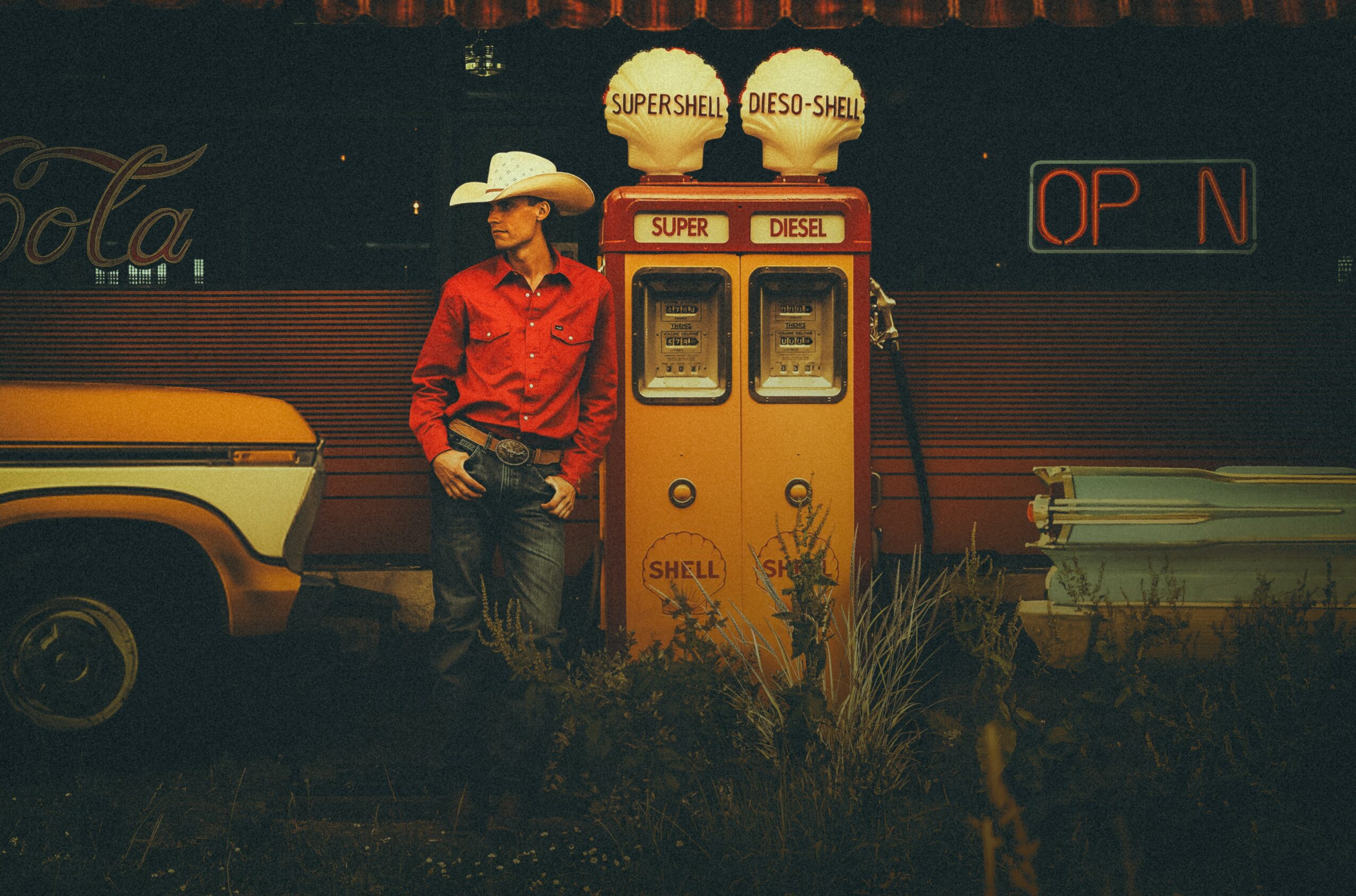 Person in red shirt and cowboy hat standing next to vintage gas pumps outdoors, with a car and a sign in the background.