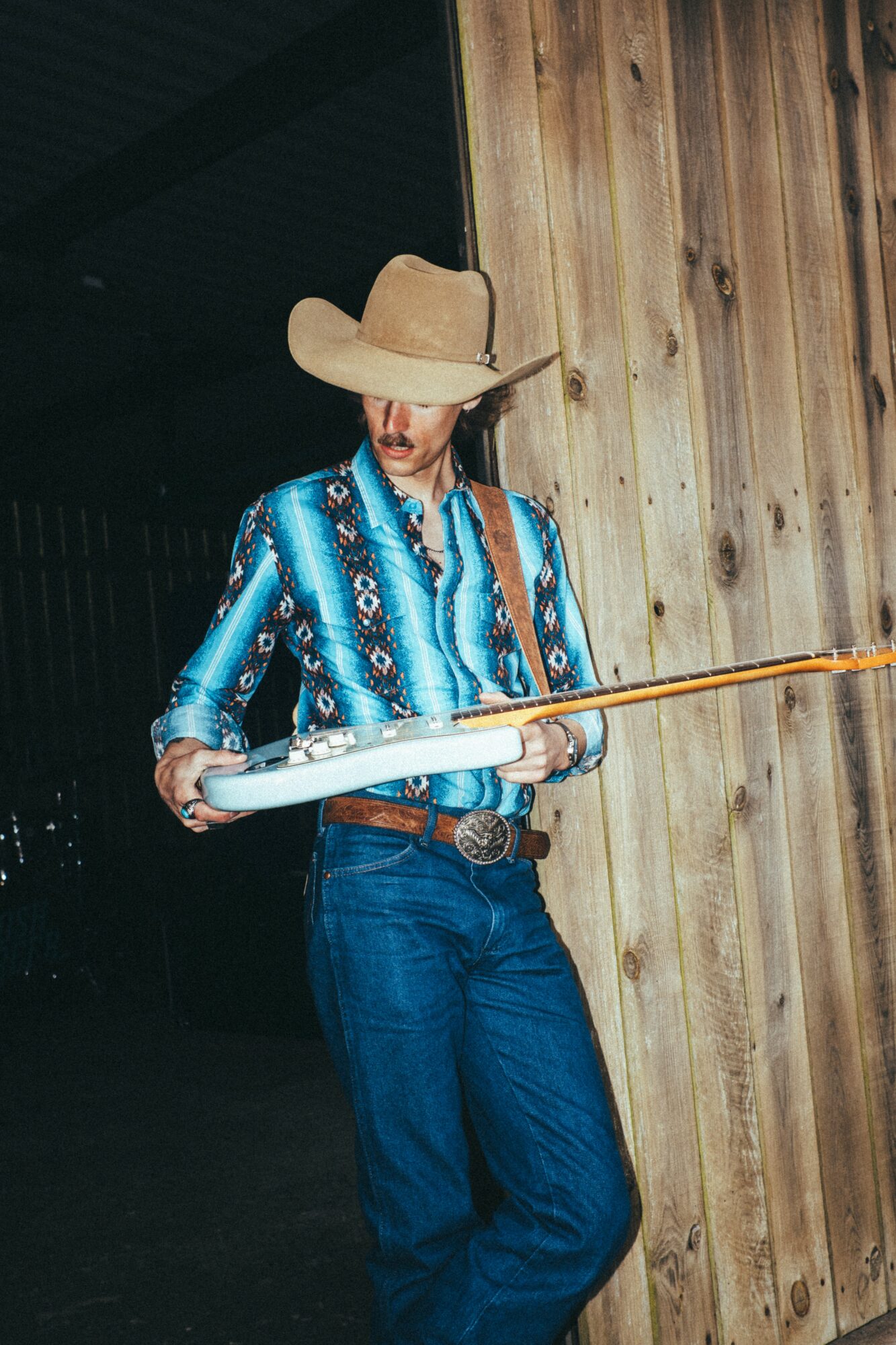 Person wearing a large hat and blue patterned shirt holding a long stick, standing against a wooden wall.