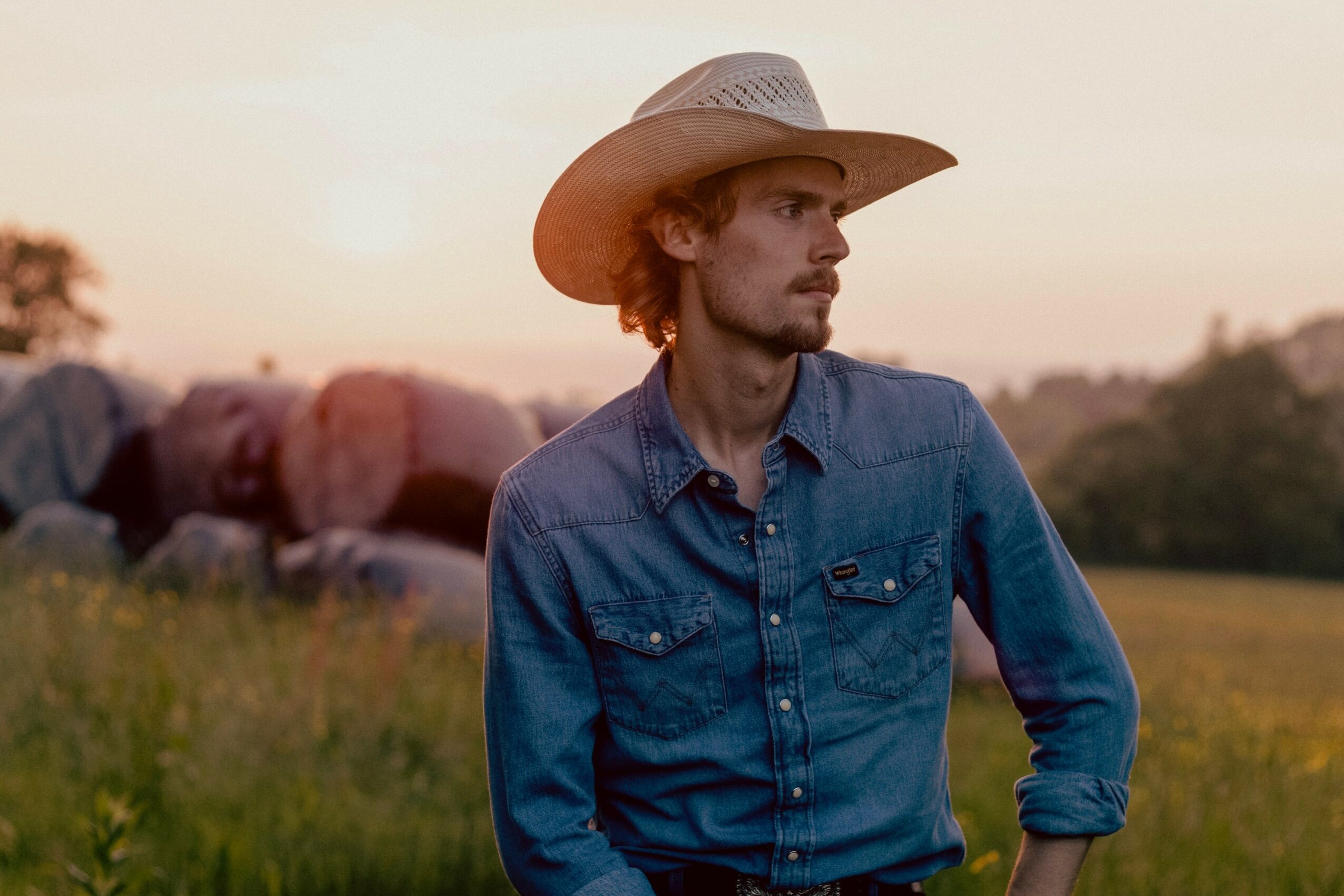 A man wearing a cowboy hat and denim shirt stands outdoors during sunset, with a field and rocks in the background.