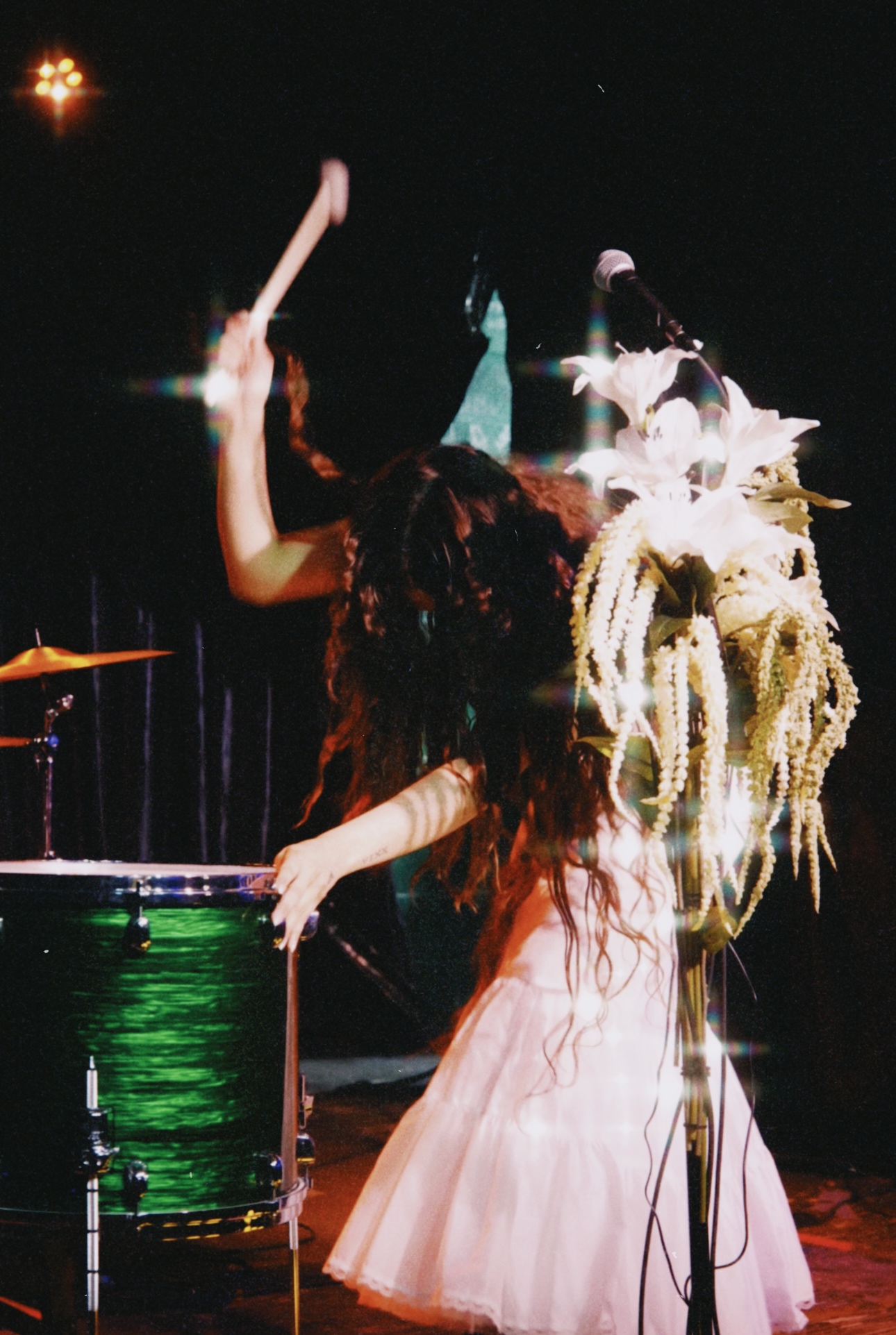 Two children with elaborate headpieces perform on stage, one playing a drum, the other in a pink dress, dark background.