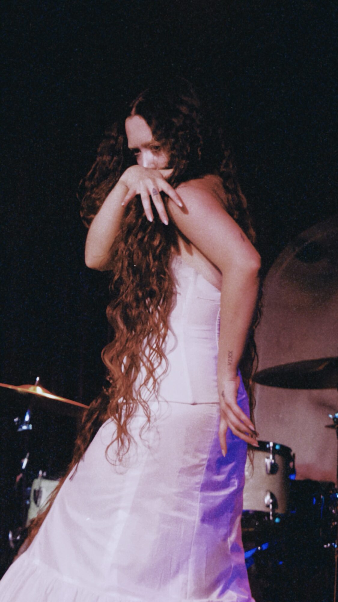 Woman with long curly hair in a white dress covering her face, standing near drums on stage.