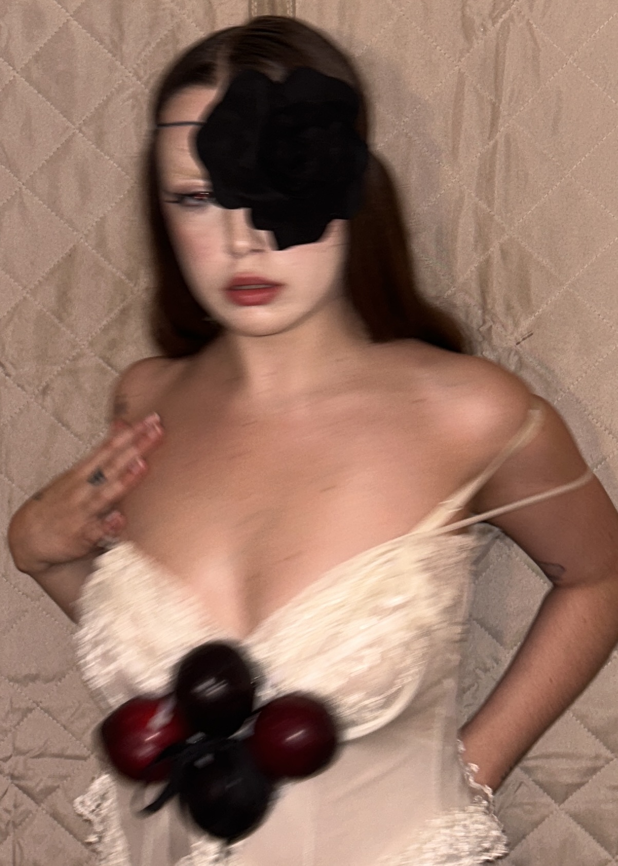 Woman with long hair wearing a white dress, holding grapes, with a black object covering her eye, standing against a quilted background.