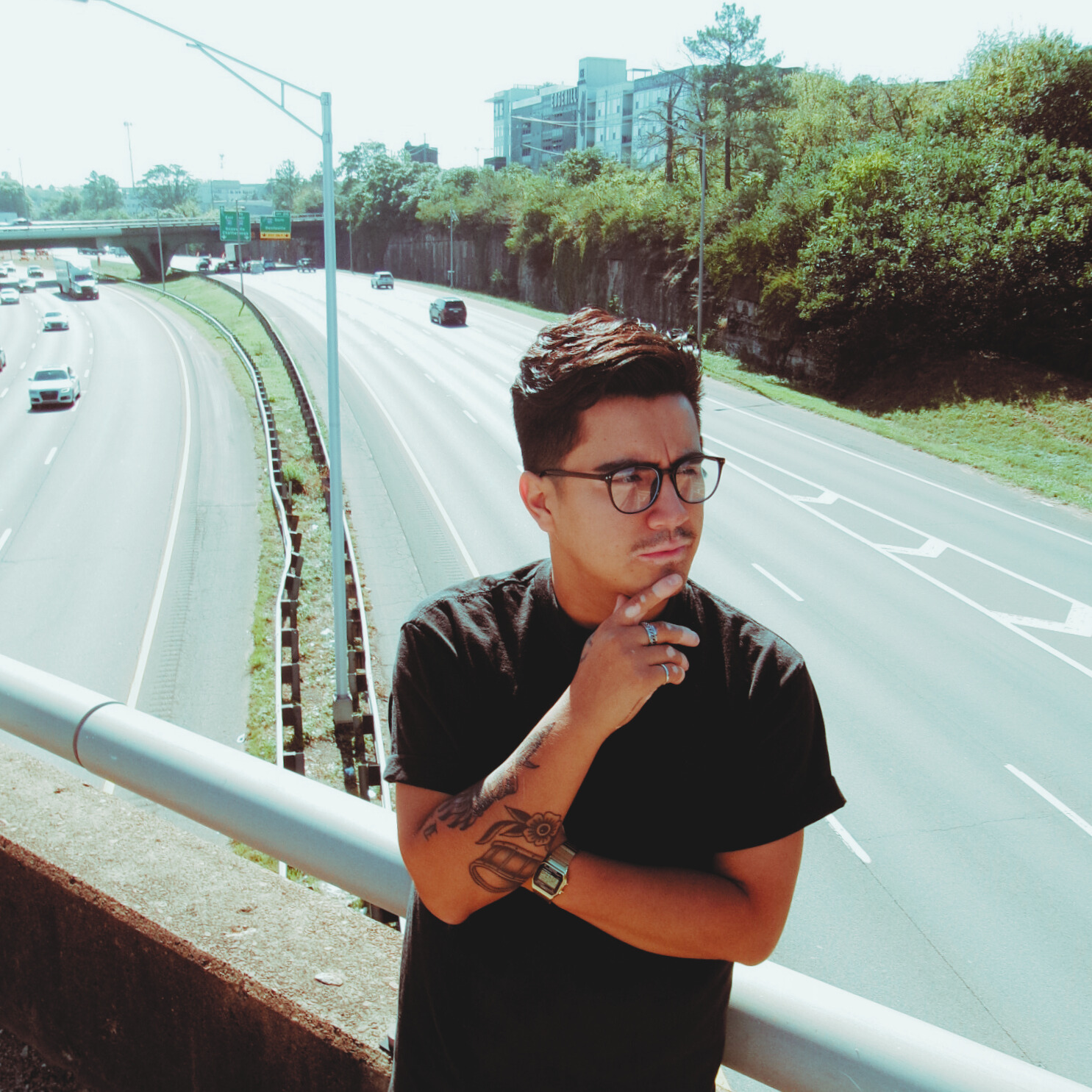Young man with glasses and tattoos standing by a highway railing, looking thoughtful, with trees and buildings in the background.
