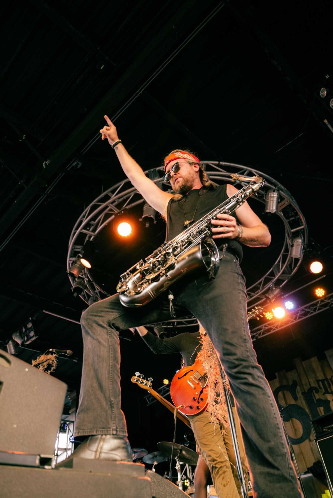 Musician with a saxophone on stage, pointing upward, wearing glasses and a black shirt, with stage lights overhead.