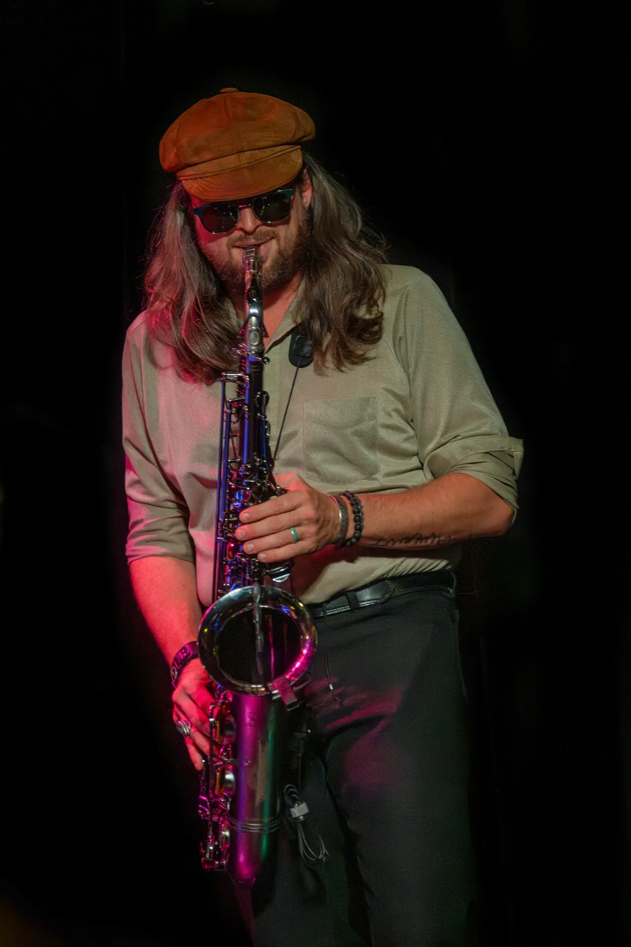 Person playing saxophone, wearing a hat and sunglasses, with long hair, against a dark background.