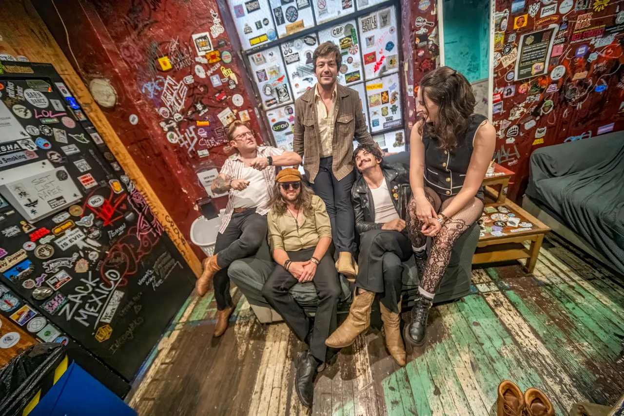 Five people sitting and standing in a colorful room with walls covered in stickers and artwork, some smiling and talking.