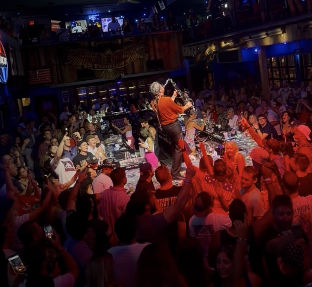 DJ performs on stage with crowd dancing in a lively nightclub, colorful lighting, and a balcony above. Word count: 20.