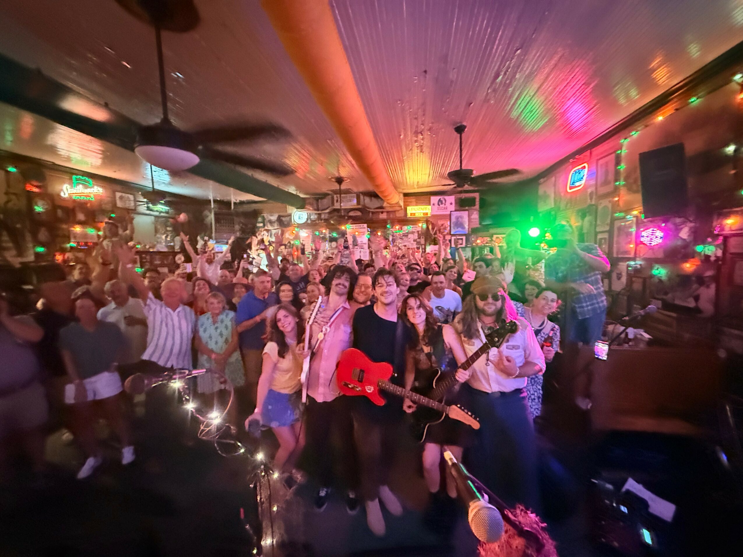 Crowd of people in a lively indoor venue with colorful lights, some holding musical instruments, celebrating together.