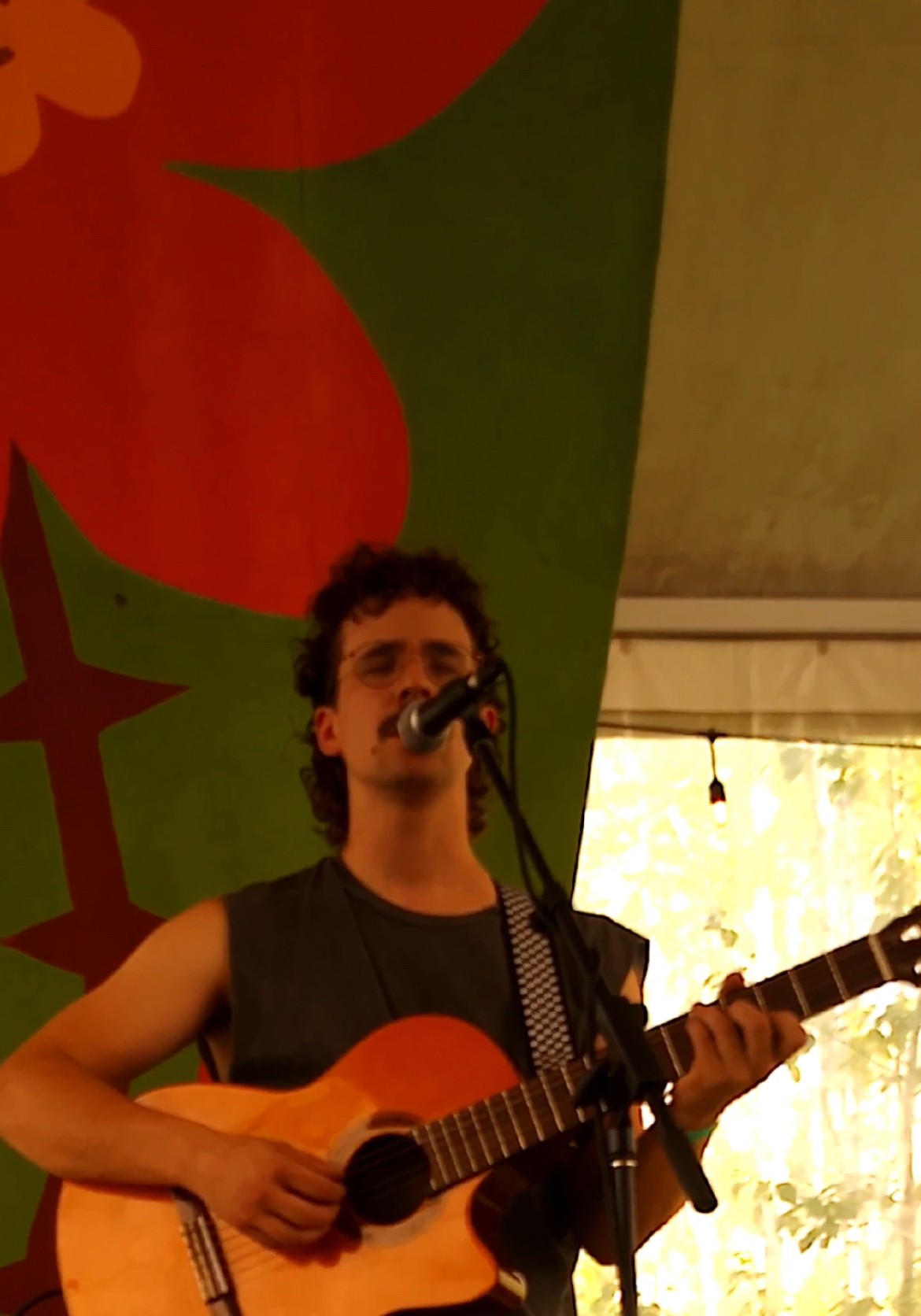 Person with curly hair playing guitar and singing into a microphone indoors, with a colorful mural in the background.