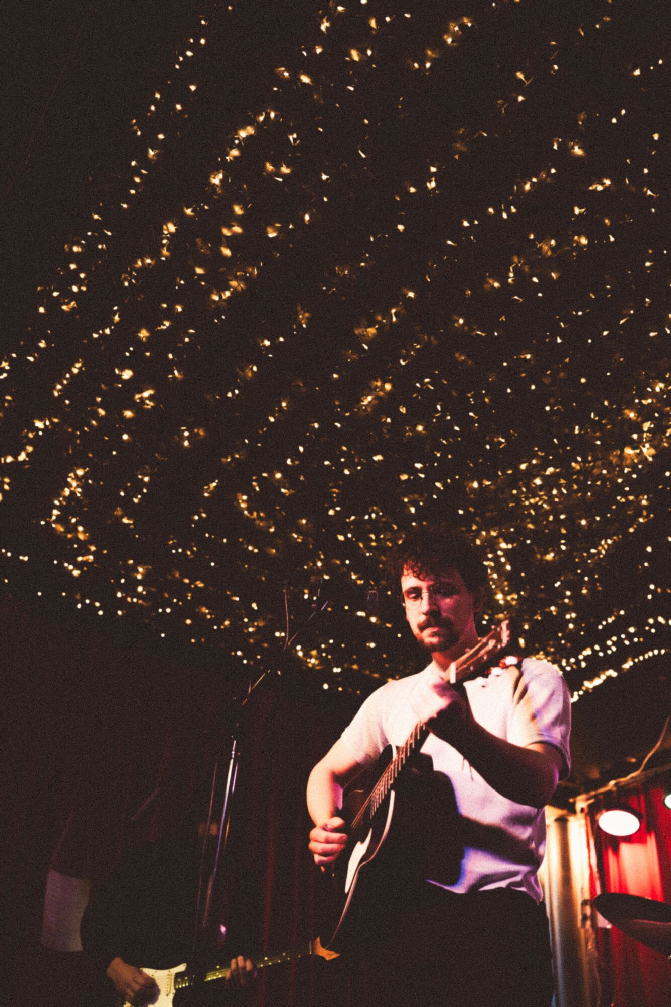 Person playing guitar on stage with a starry ceiling background, dim lighting, and red curtains.