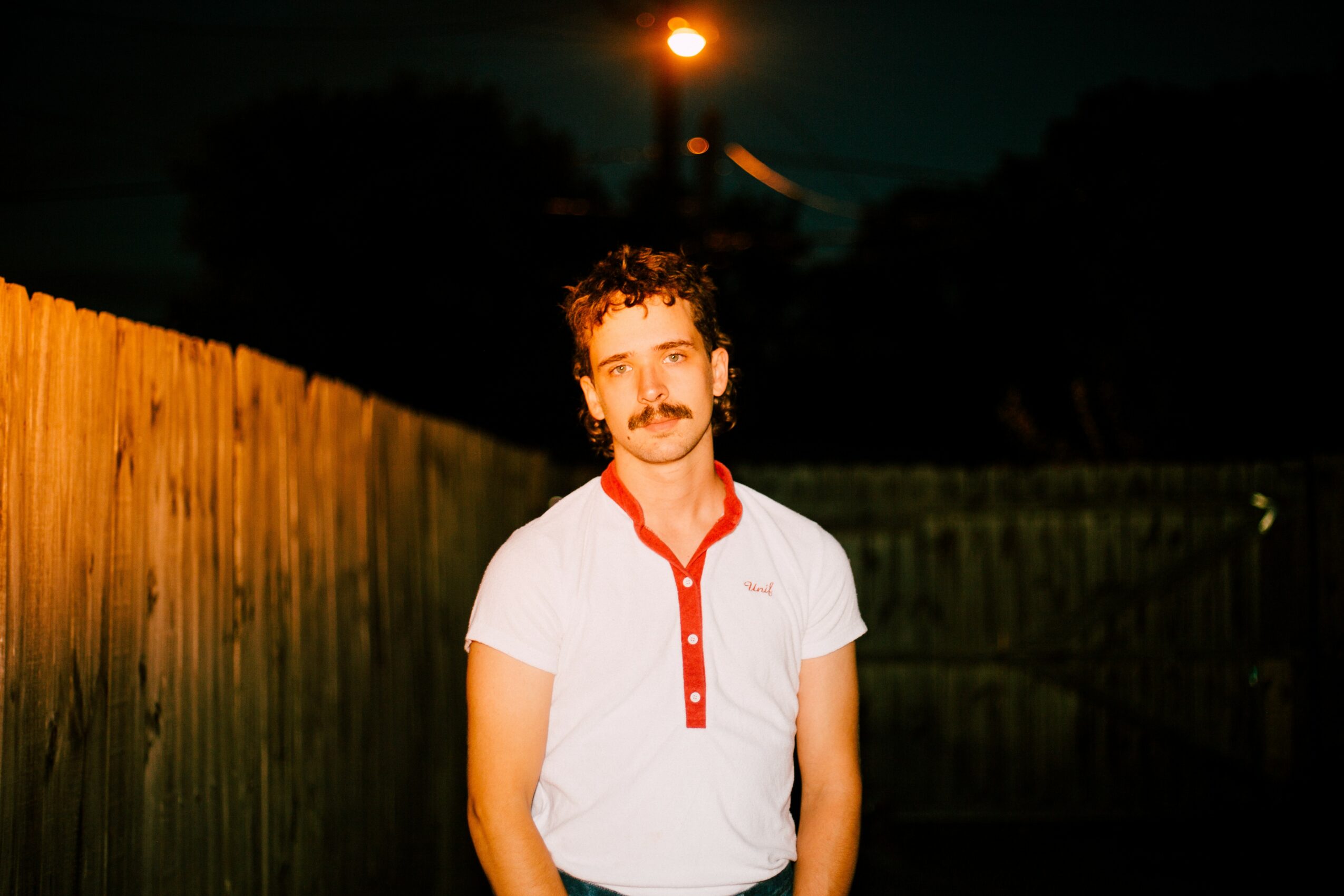Young man with curly hair and mustache wearing a white shirt with red buttons, standing outdoors at night.