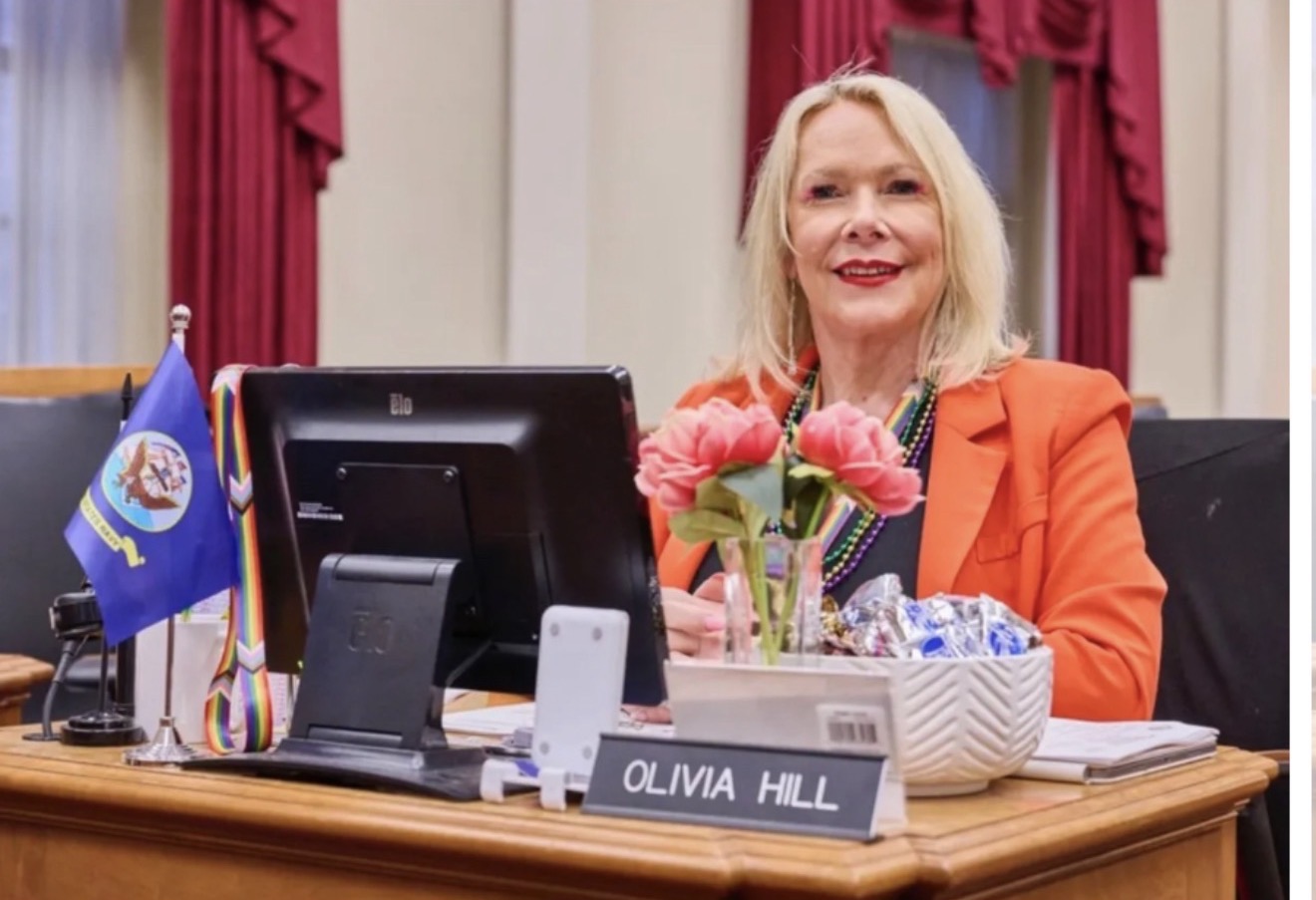 Woman sitting at a desk with a computer, flowers, and flags, smiling in a formal setting.