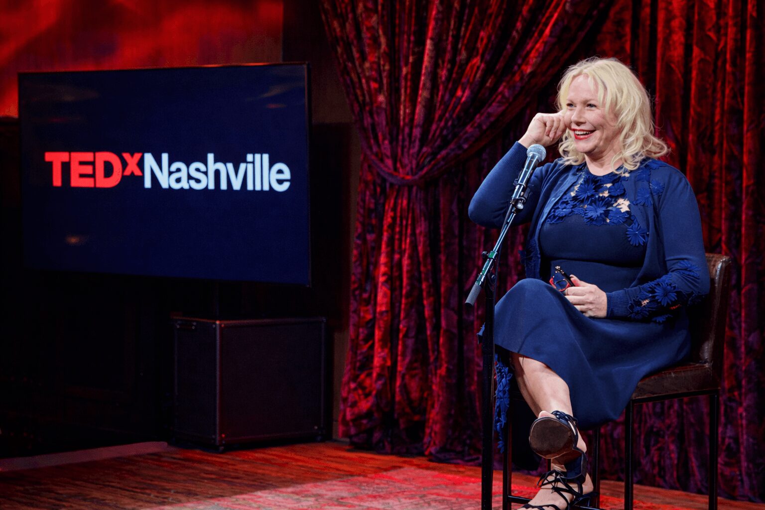 Woman sitting on a chair, smiling, holding a phone, with a TEDx Nashville sign and red curtain background.